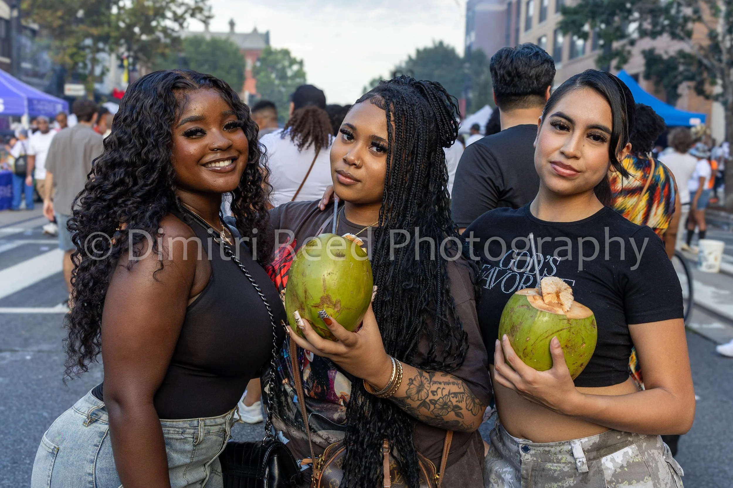 Three women holding coconuts with straws at an outdoor event, surrounded by a crowd, with tents and trees in the background.