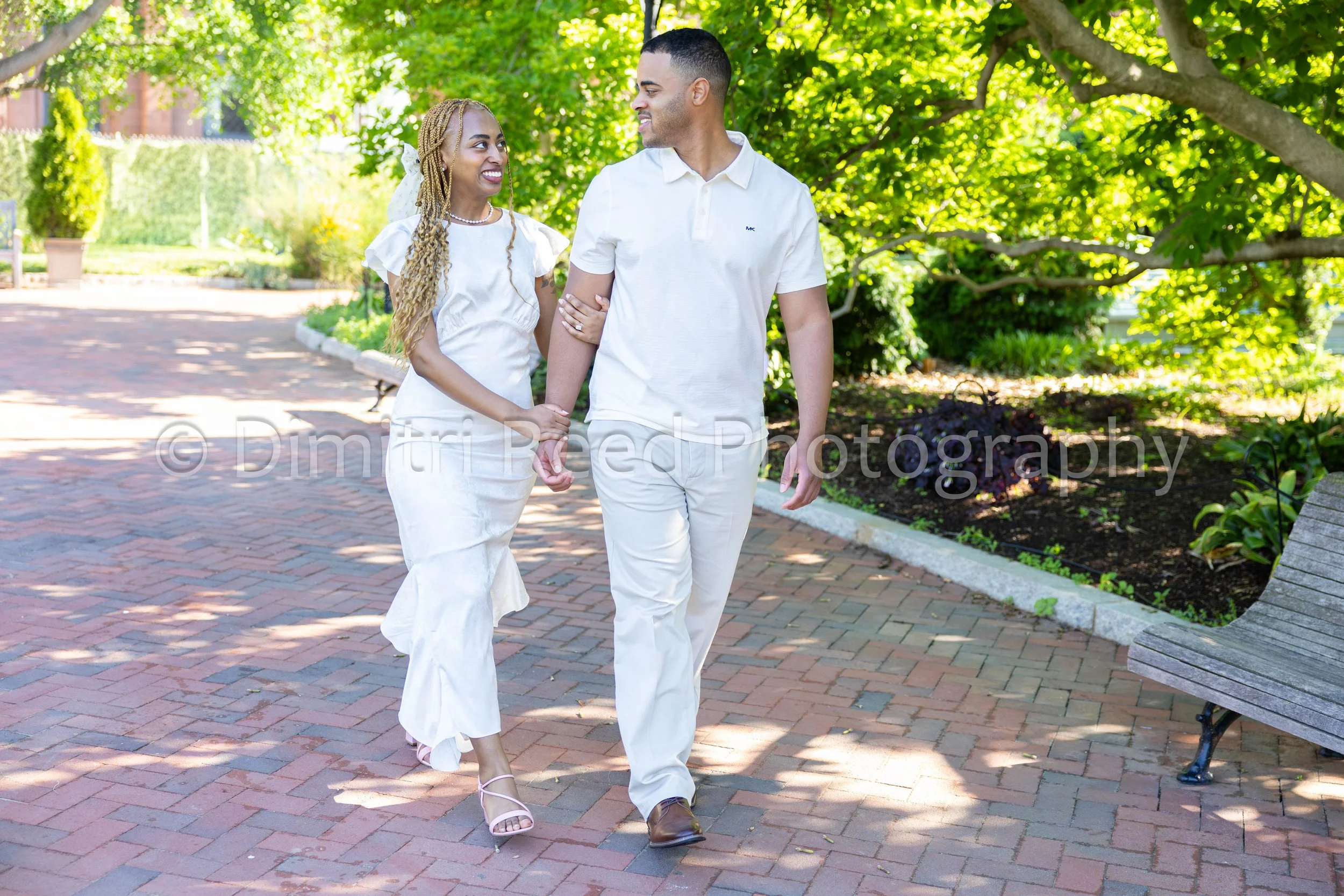 A couple walking hand in hand in a park, smiling and looking at each other, surrounded by green trees and sunlight.