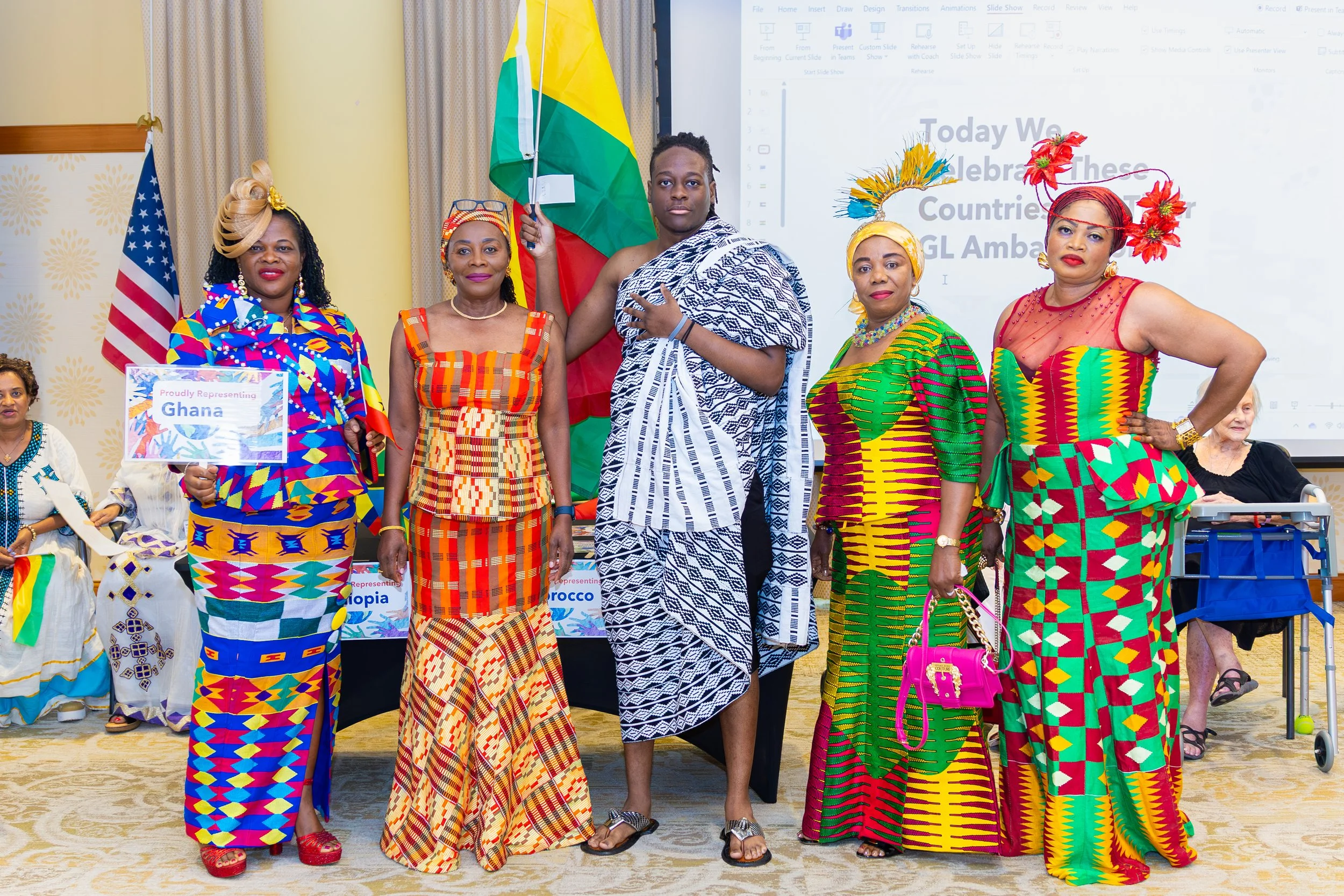 Group of five women and one man, dressed in colorful traditional African attire, standing indoors at a cultural celebration event. The man is holding the flag of Guyana. Two women are holding signs representing Ghana and Ethiopia. In the background, 