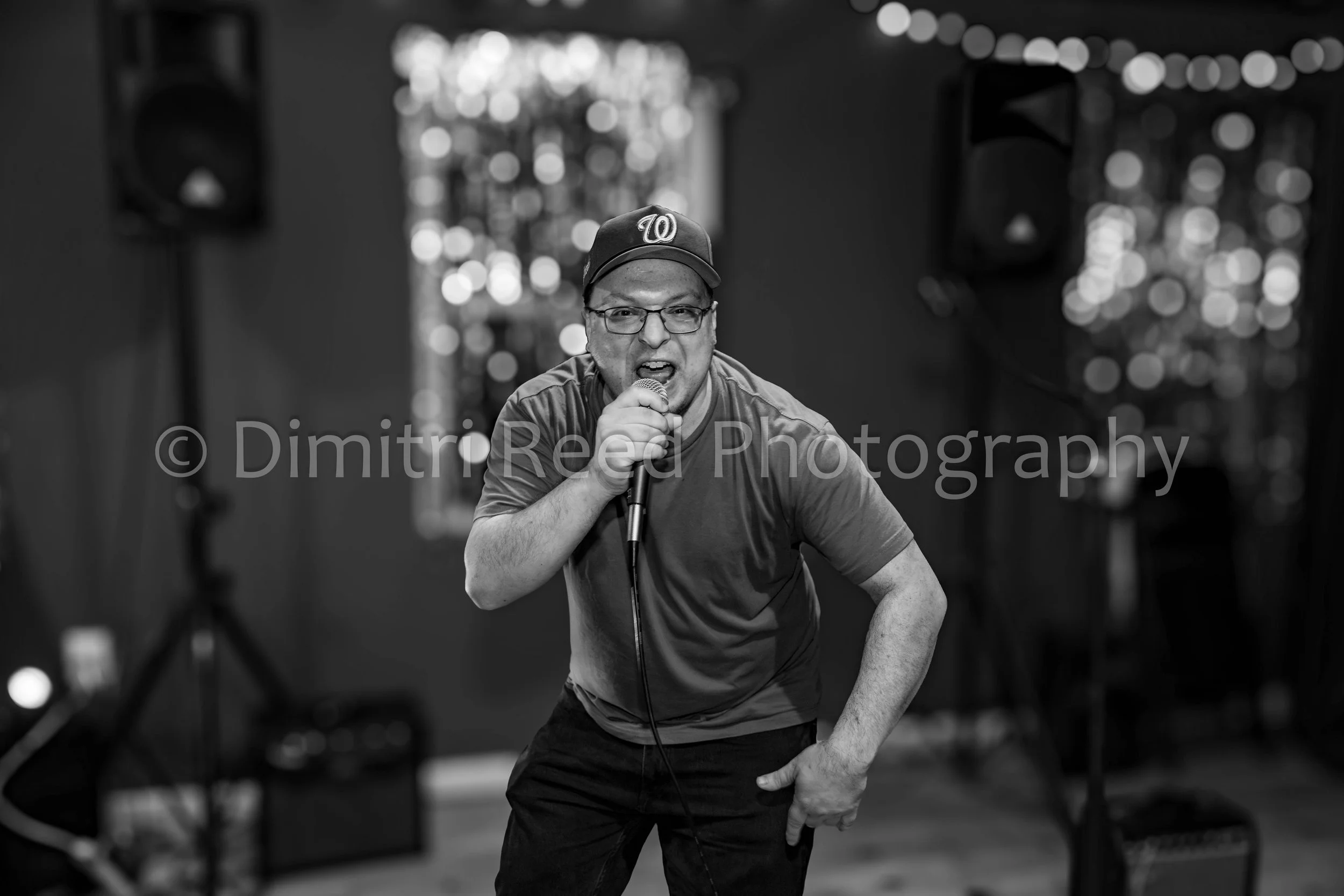 A man wearing glasses, a cap with a 'W' logo, and a t-shirt, appears to be speaking or singing into a microphone on a stage or performance space. The background is blurred with bokeh lights.
