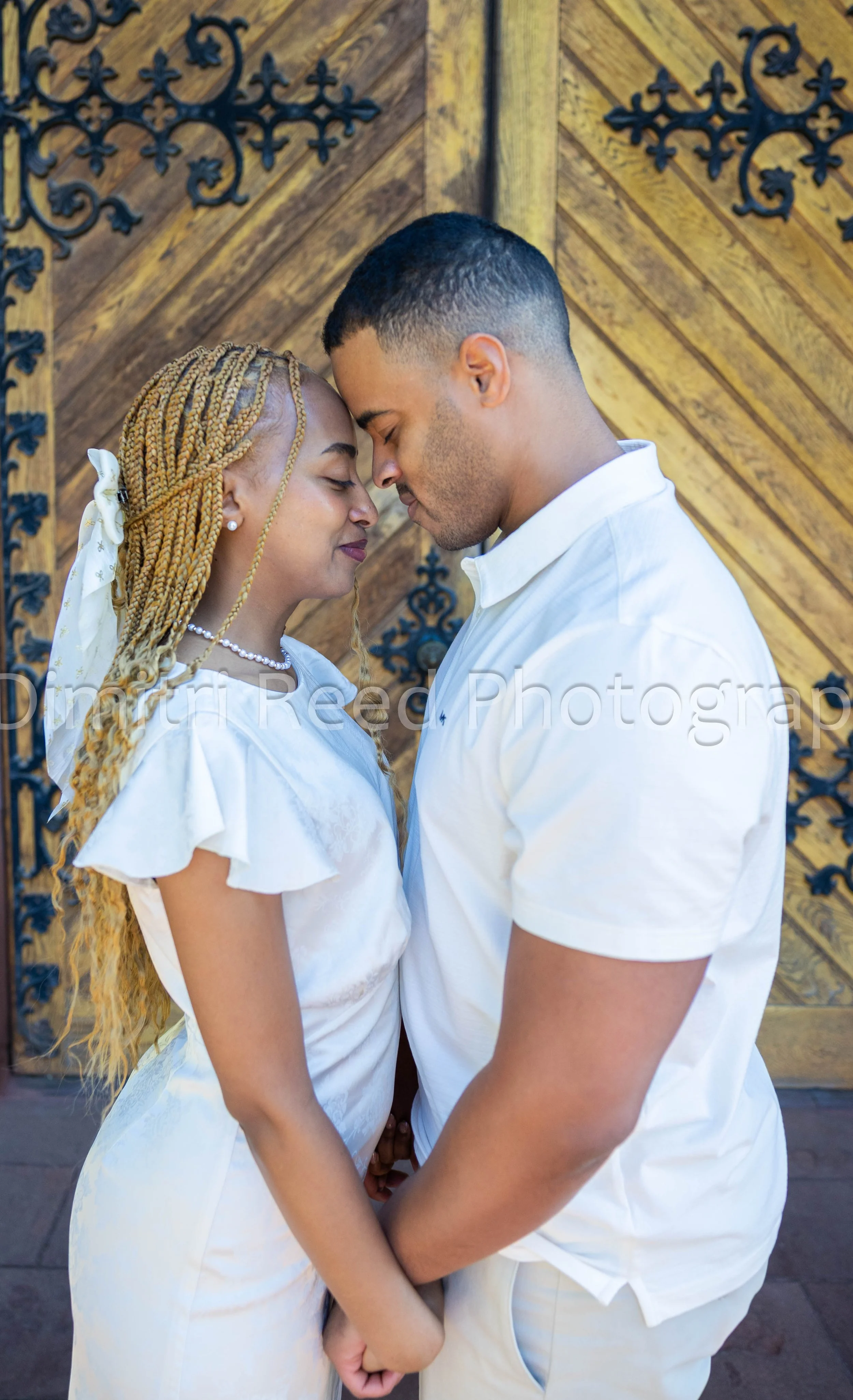 A couple with foreheads touching, eyes closed, holding hands, standing in front of a wooden door with decorative black ironwork.