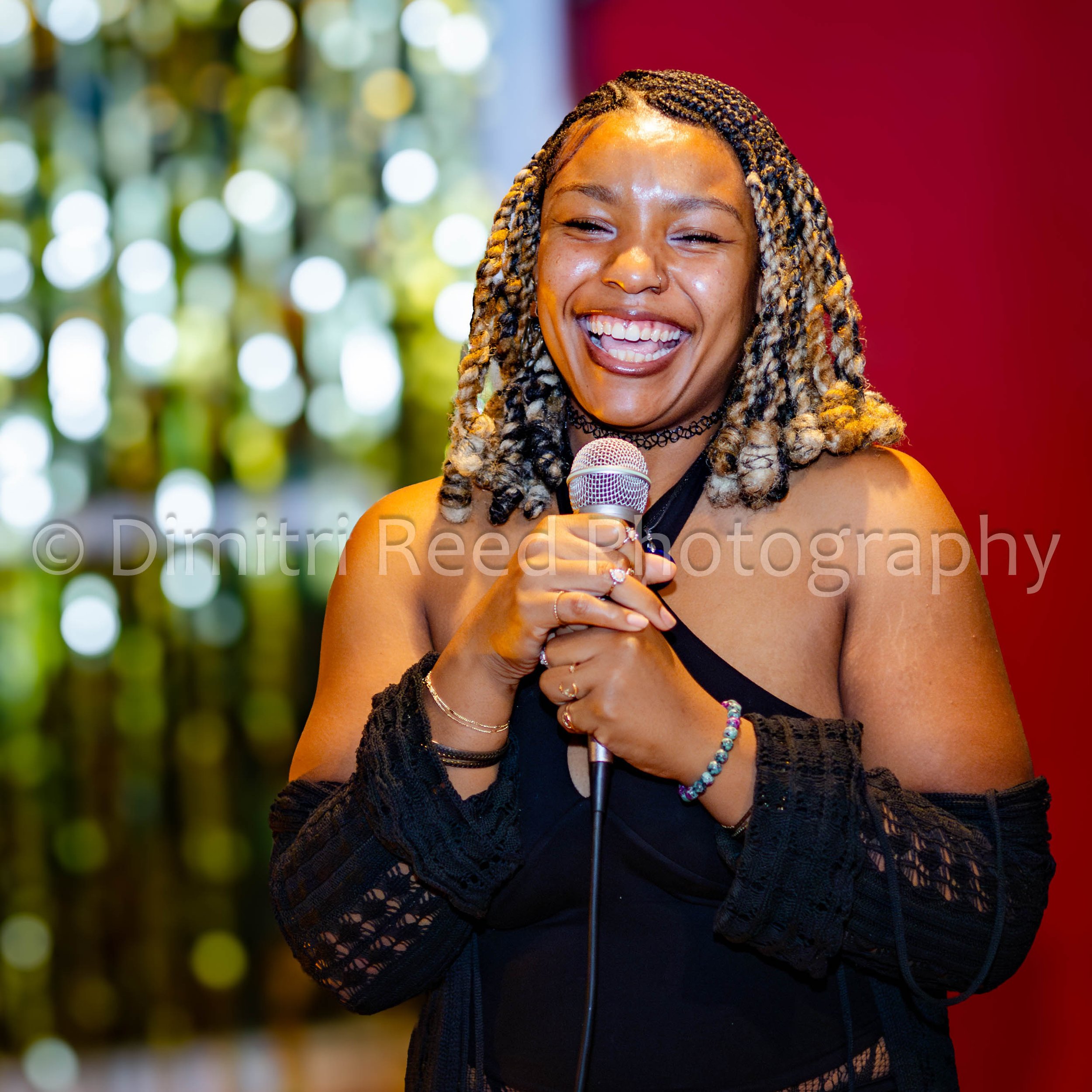 A woman with braided hair smiling and holding a microphone, standing in front of a colorful background with bokeh lights and red wall.
