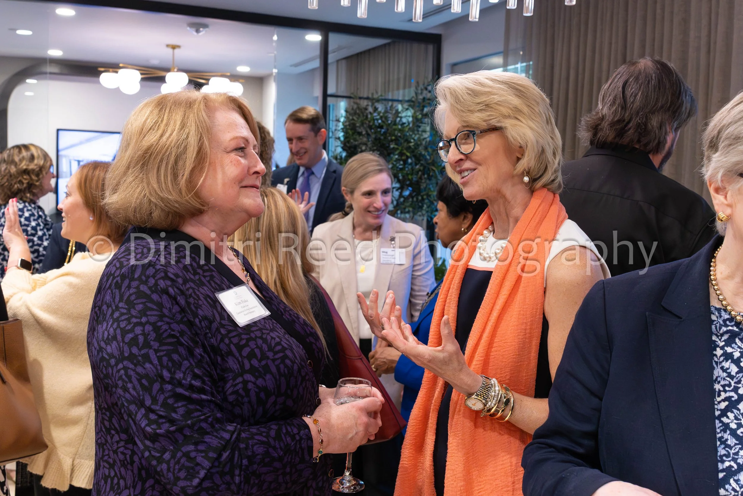 Two women engaged in conversation at a social gathering, surrounded by other attendees in a brightly lit indoor space.