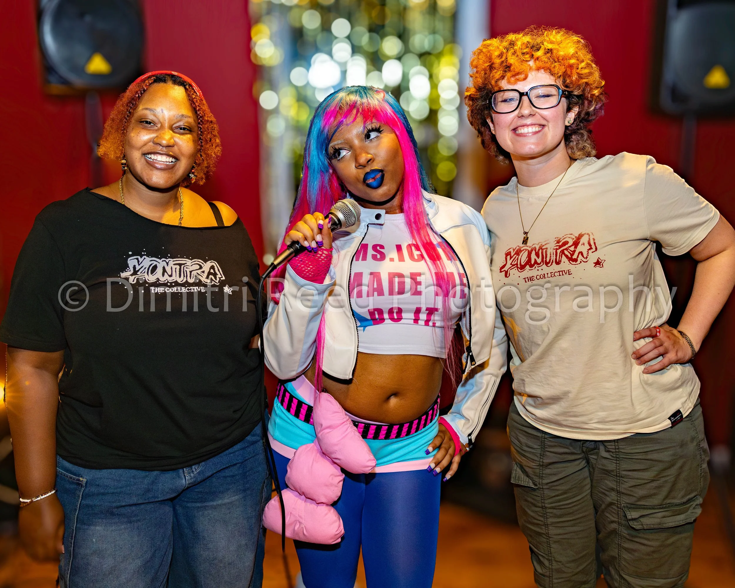 Three women standing together and smiling, one with colorful hair and makeup holding a microphone, in a setting with blurry lights and red walls.