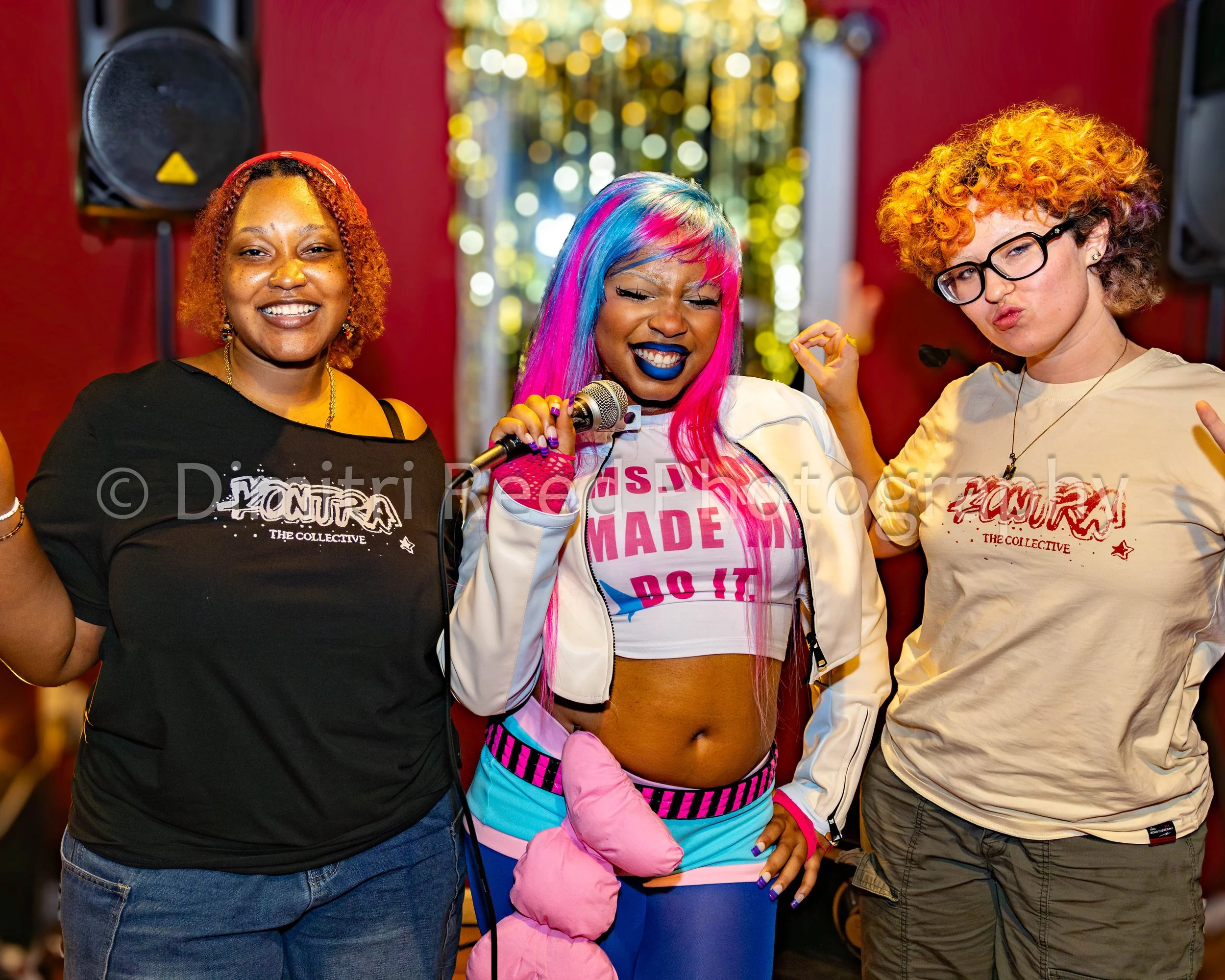 Three women standing together in a festive indoor space with colorful lights in the background. The woman in the middle has multi-colored hair, blue lipstick, and is dressed in athletic shorts, a white cropped hoodie, and pink gloves, smiling and hol