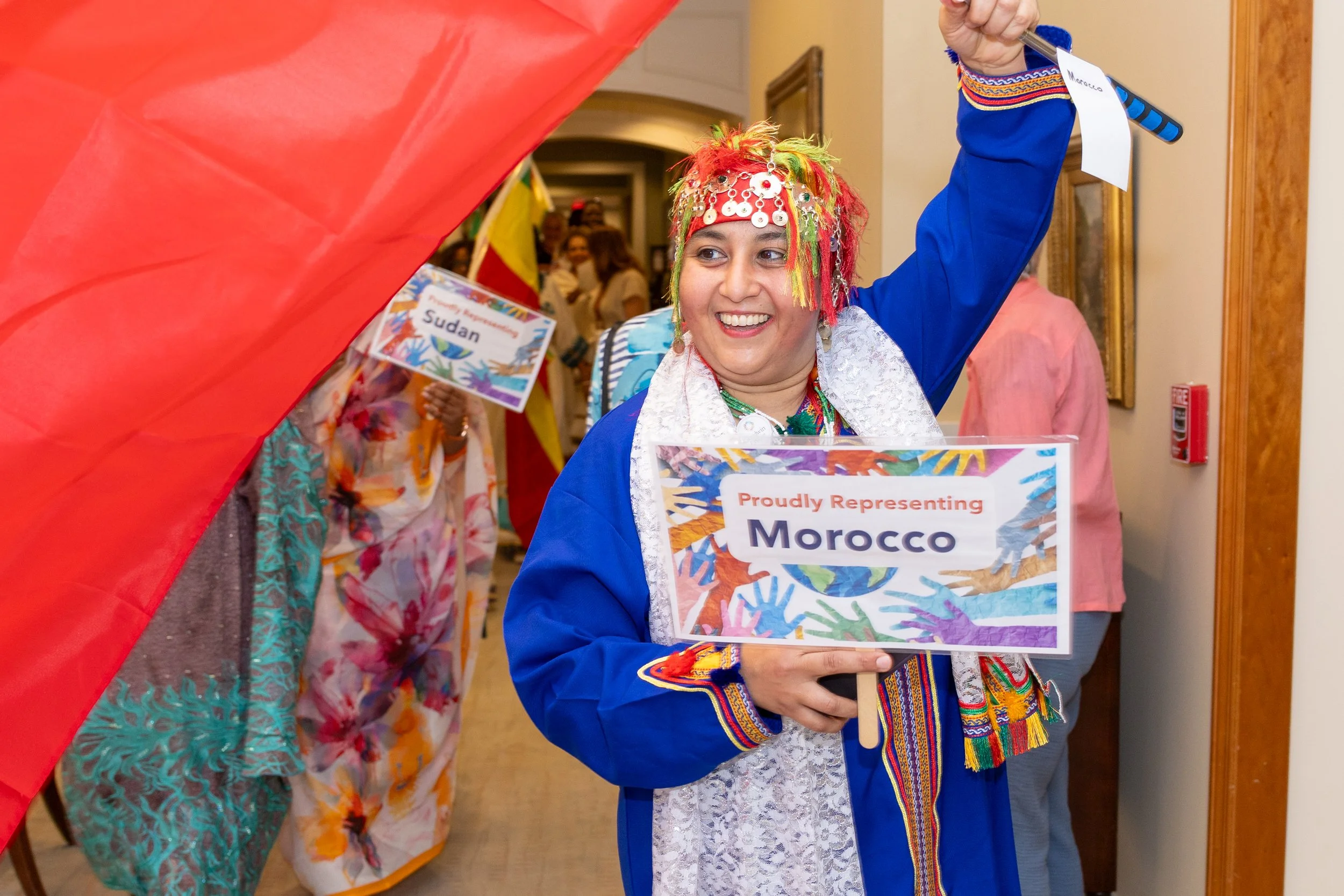 A woman in colorful, traditional Moroccan attire is smiling and holding a sign that says 'Proudly Representing Morocco'. She is surrounded by other people participating in what appears to be a parade or cultural event. The woman is wearing a red head
