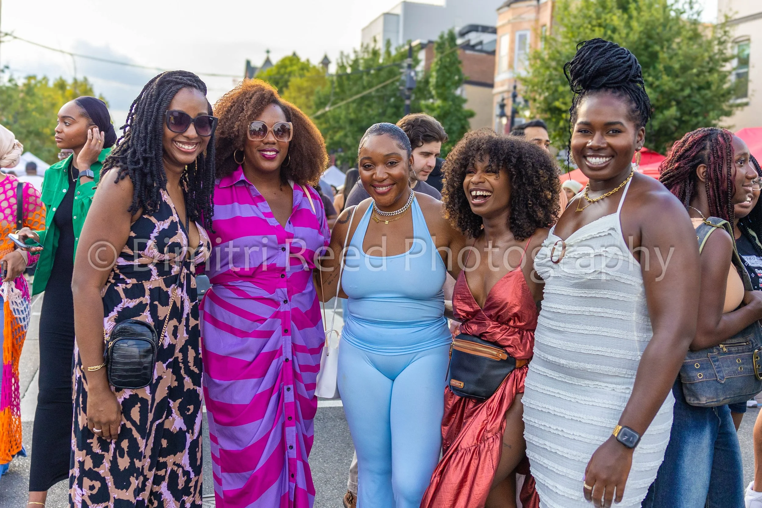 A group of women at an outdoor event smiling and standing close together, dressed in colorful outfits, with trees and buildings in the background.