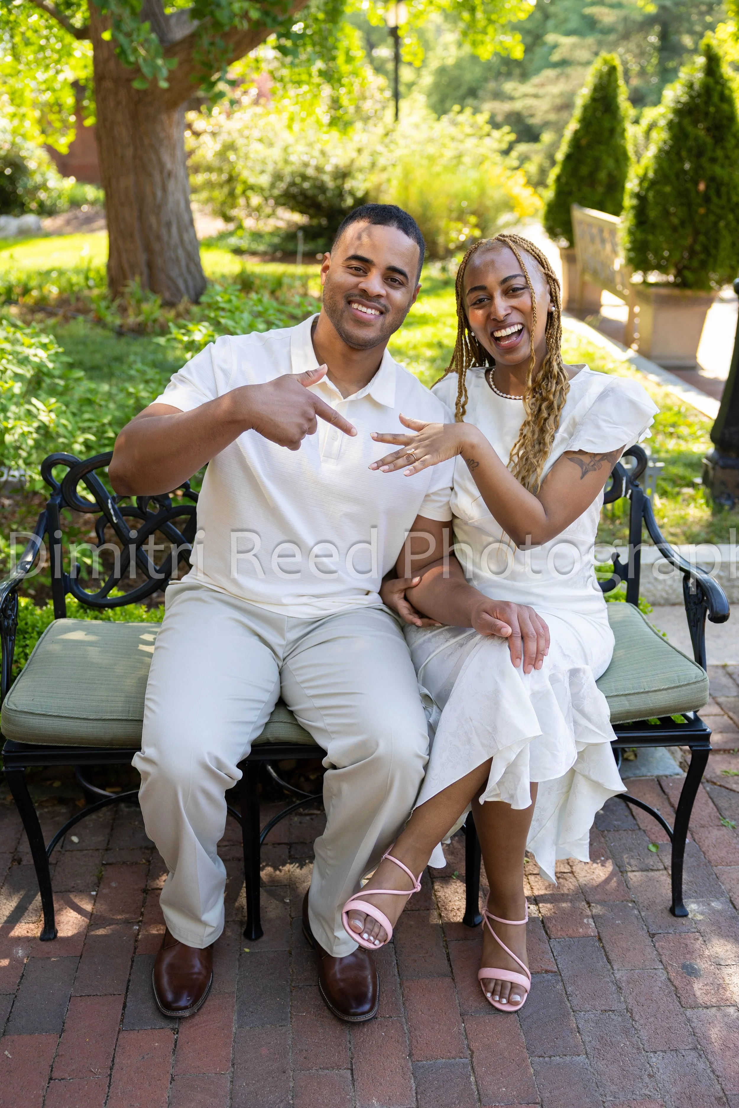 A smiling man and woman sitting on a park bench outdoors, showing off the woman's engagement ring. The man is pointing at the ring, and they are both celebrating. The background features trees, bushes, and a sunny, green park scene.