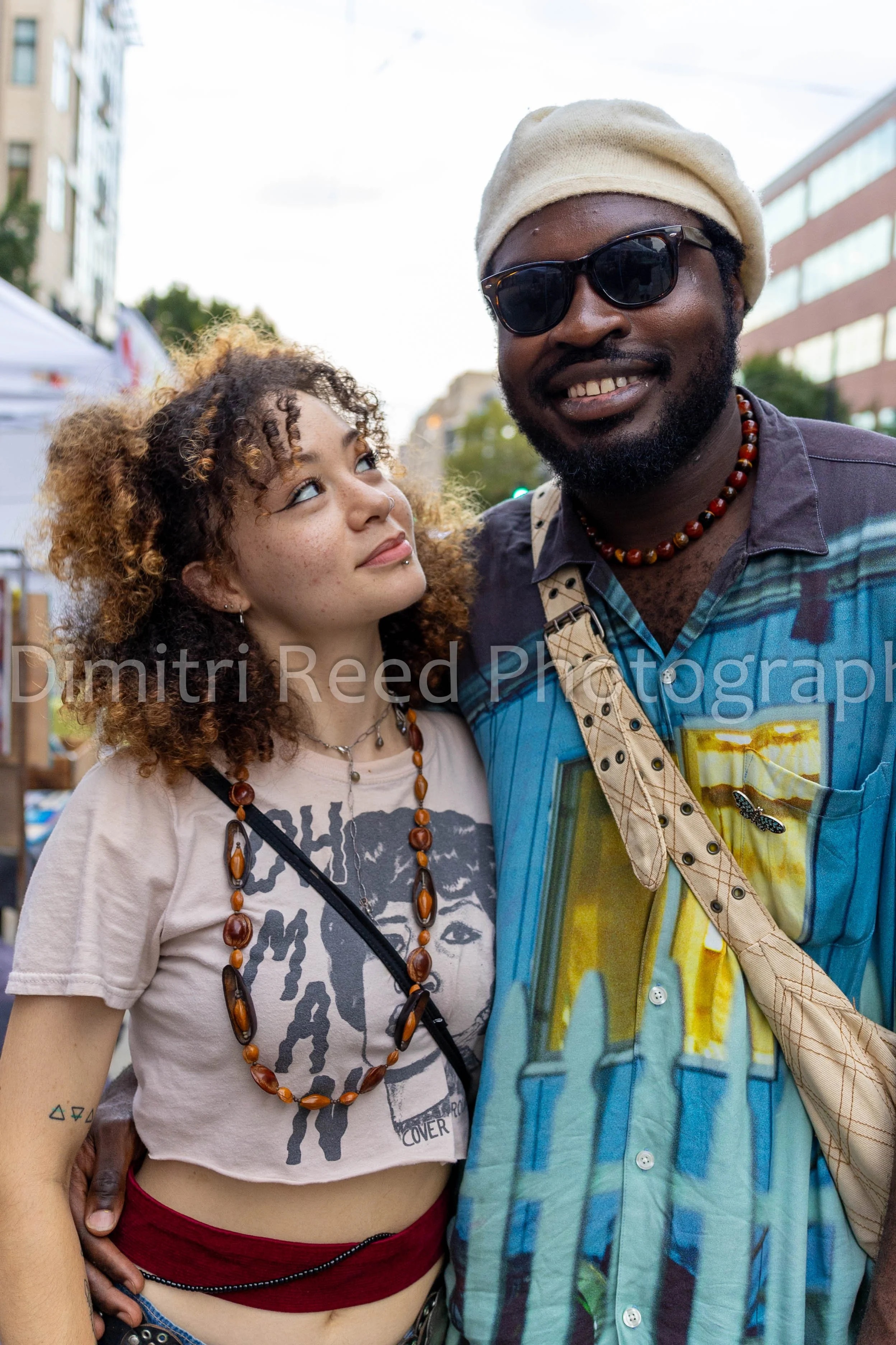 A young woman with curly hair, wearing a graphic t-shirt, red pants, and a large amber beaded necklace, standing close to a bearded man wearing sunglasses, a cream hat, a colorful patterned shirt, and a necklace, outdoors in an urban setting with bui