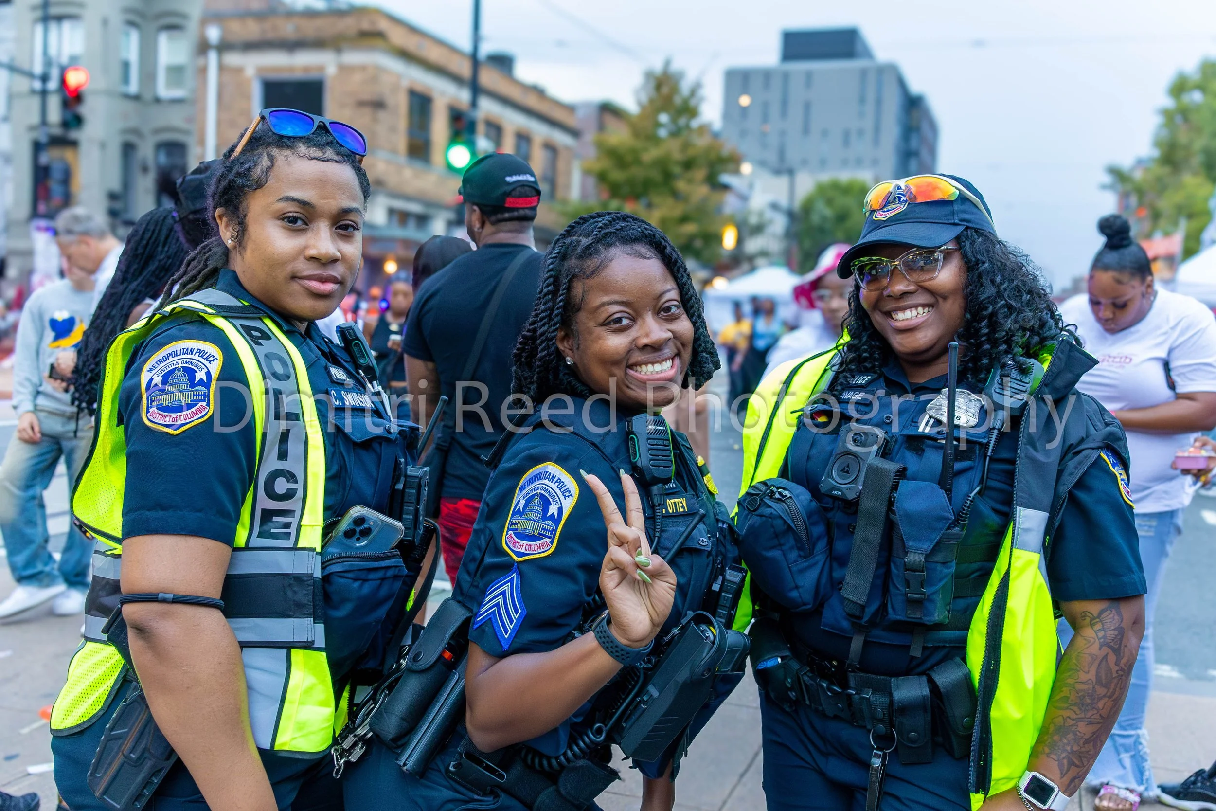 Three female police officers standing together outdoors in an urban setting, smiling and posing for the camera. They are in uniform with police badges, and the background shows a crowd, trees, and buildings.