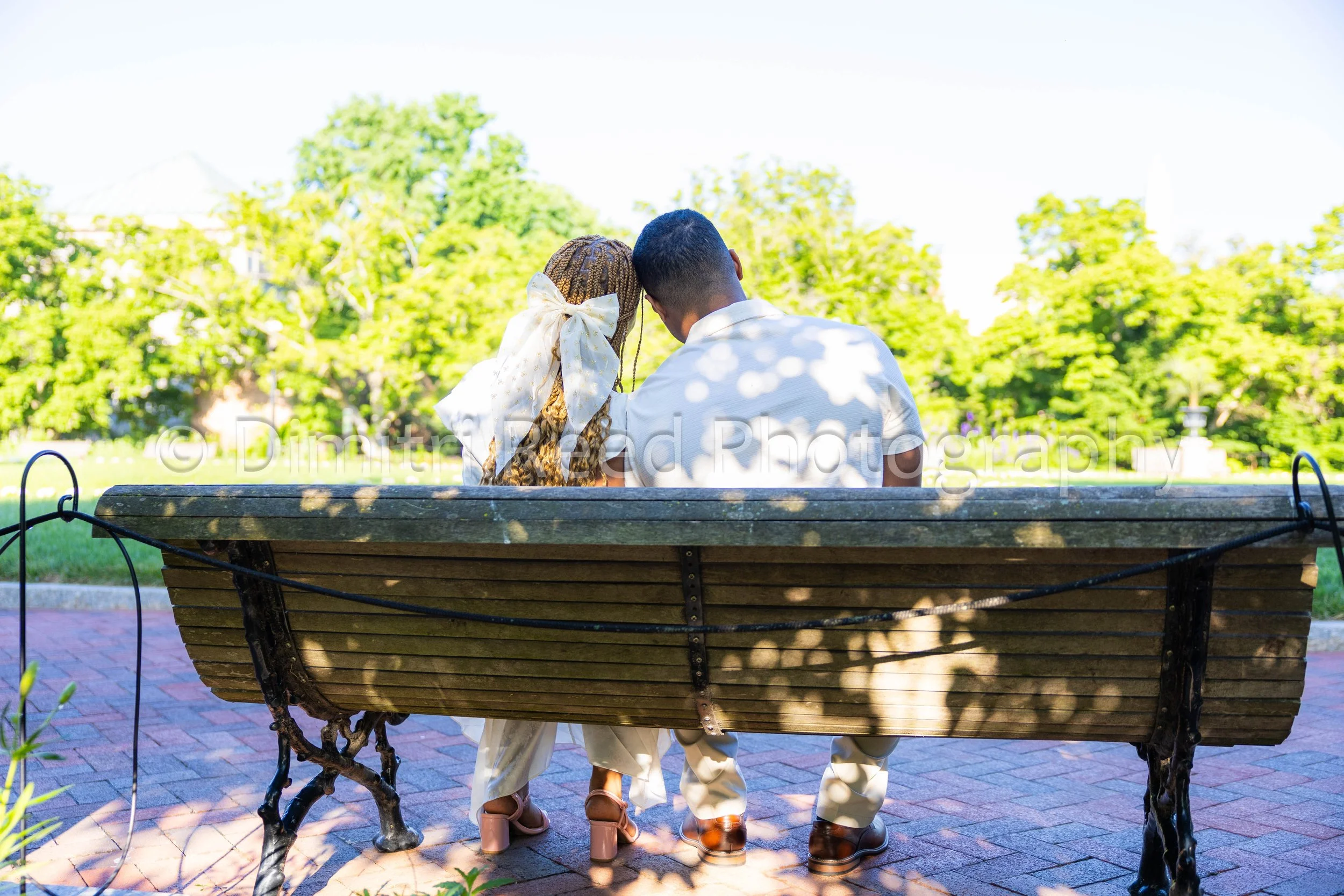 Couple sitting on a park bench under tree shadows, facing away, with lush green trees and bright sky in the background.
