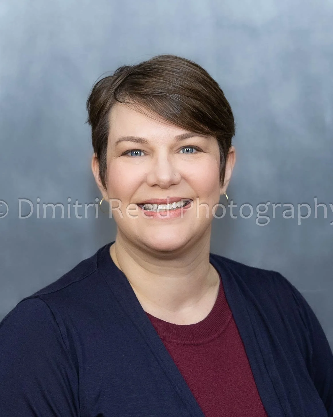 Close-up portrait of a woman with short brown hair, blue eyes, wearing a black jacket and maroon shirt, smiling against a gray background.