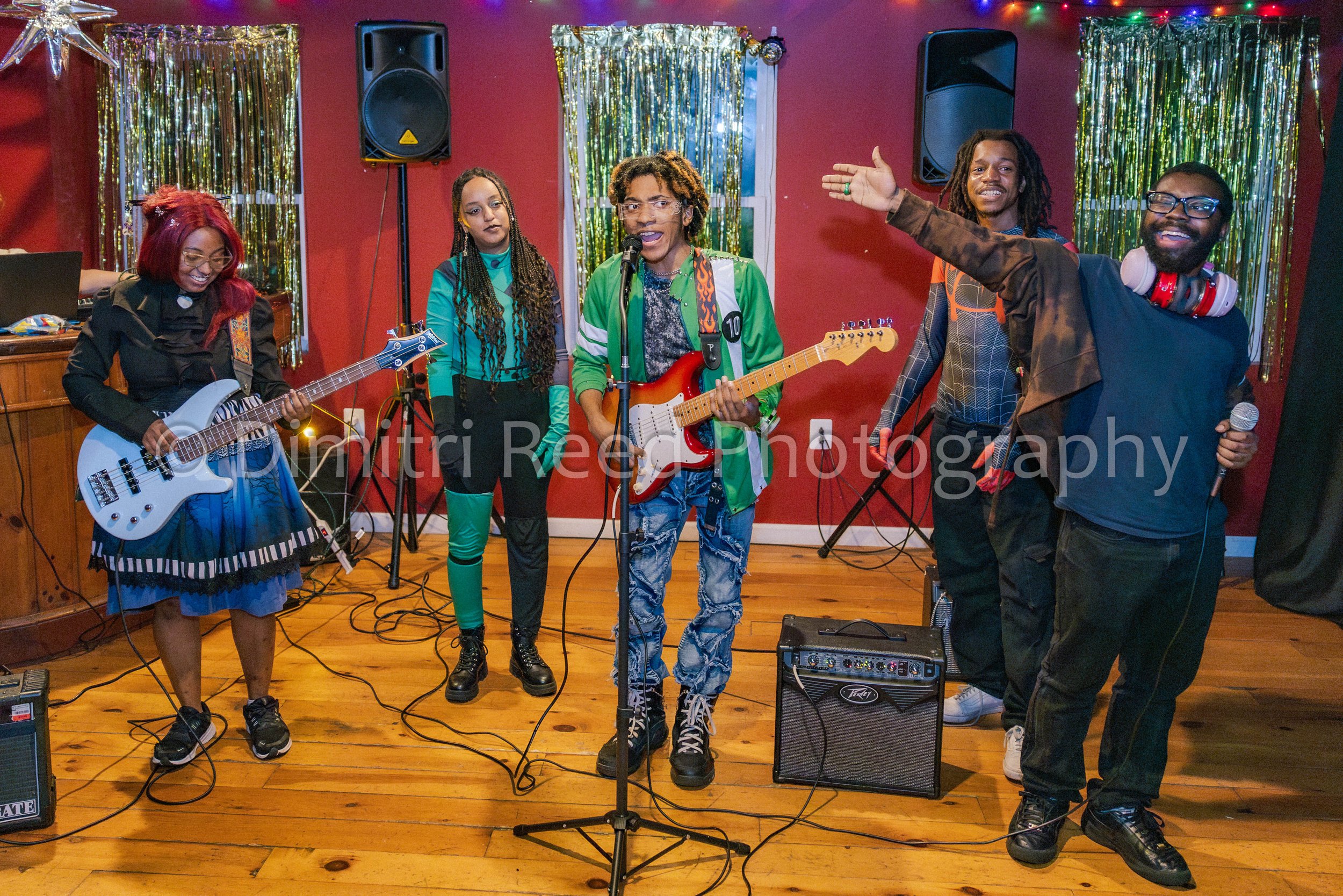 A diverse group of six people performing music together in a room decorated with gold tinsel and colorful string lights. One woman with red hair is playing a white electric bass, another woman with braids is standing in front of a microphone, the thi
