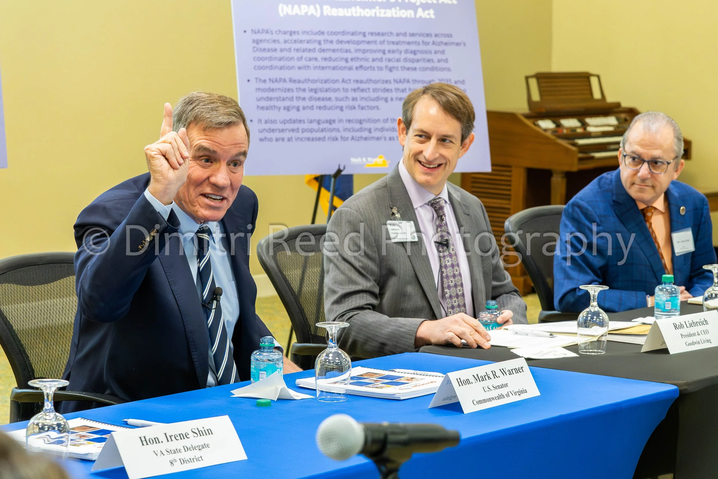Three men seated at a conference table during a meeting or discussion. The man on the left is speaking and gesturing with his right hand. The man in the middle is smiling, and the man on the right is wearing glasses and looking attentively. Nameplate