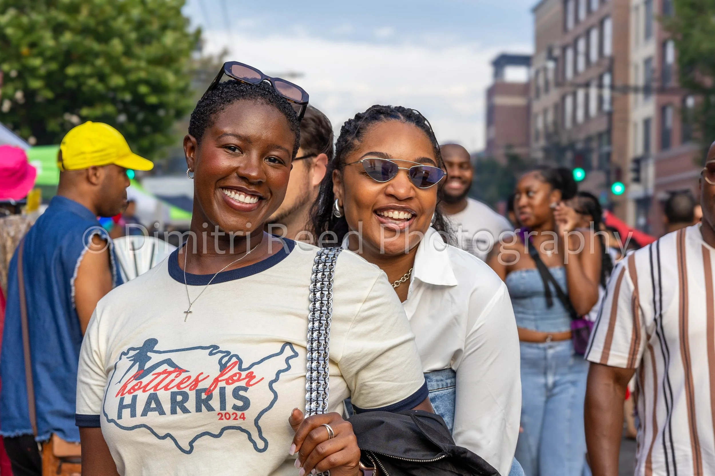 Two smiling women at an outdoor gathering, one with short curly hair and sunglasses, wearing a shirt with a political message supporting Bernie Harris 2024, and the other with braided hair and sunglasses, surrounded by other people in casual clothing