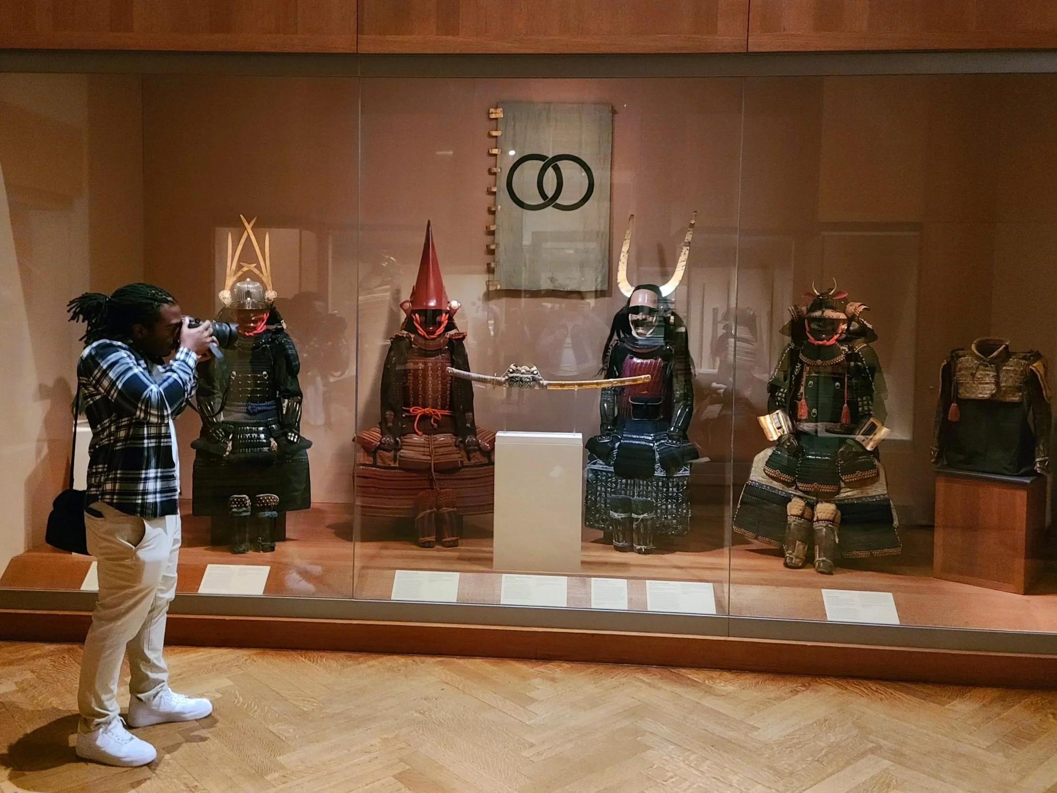 A person with dreadlocks wearing a black and white plaid shirt takes a photo of a display case showcasing traditional Japanese samurai armor and helmets in a museum. The display includes various armor pieces and a framed symbol on the wall.