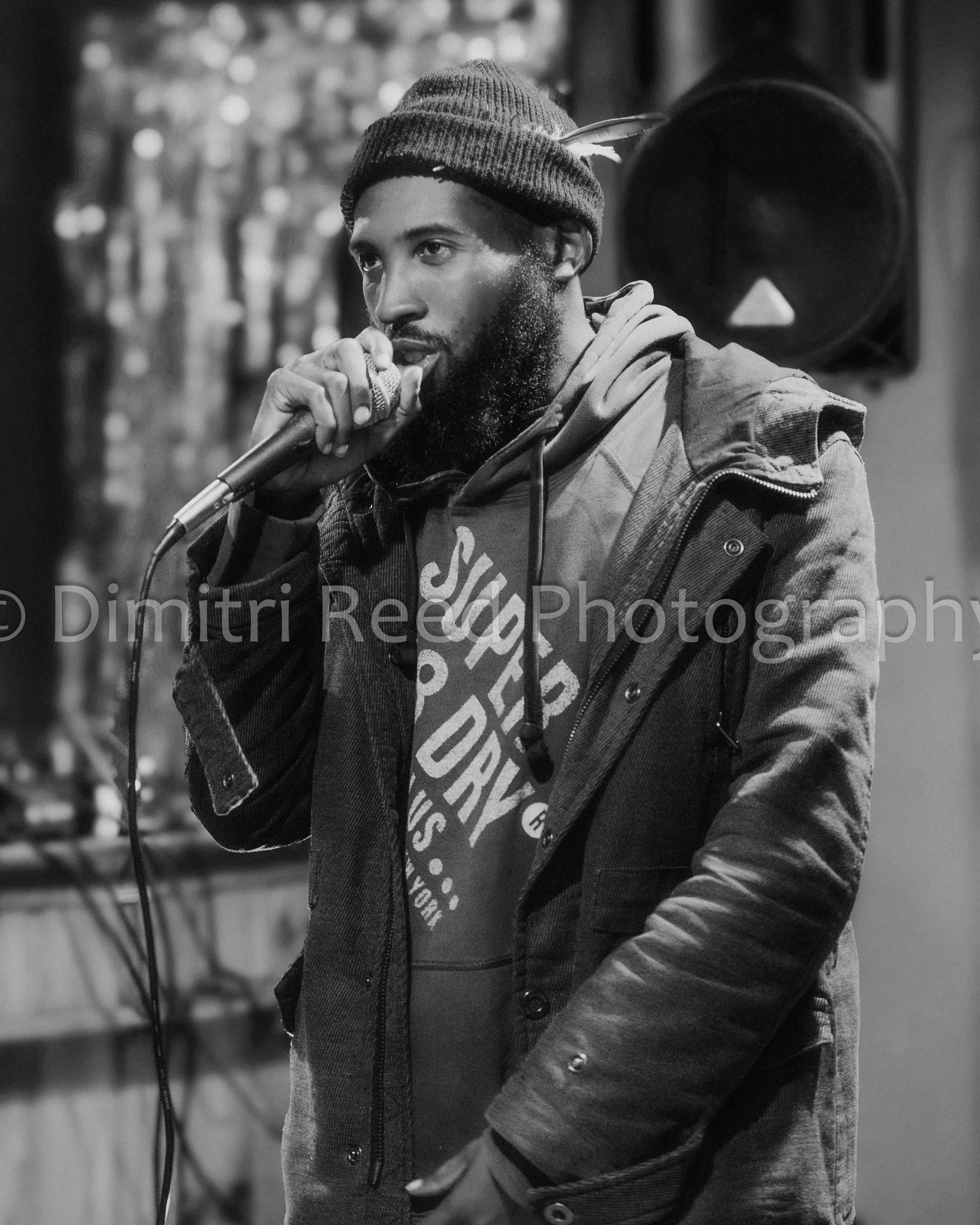 A young man singing into a microphone in a recording studio, wearing a beanie, a hoodie, and a jacket, with a speaker in the background.
