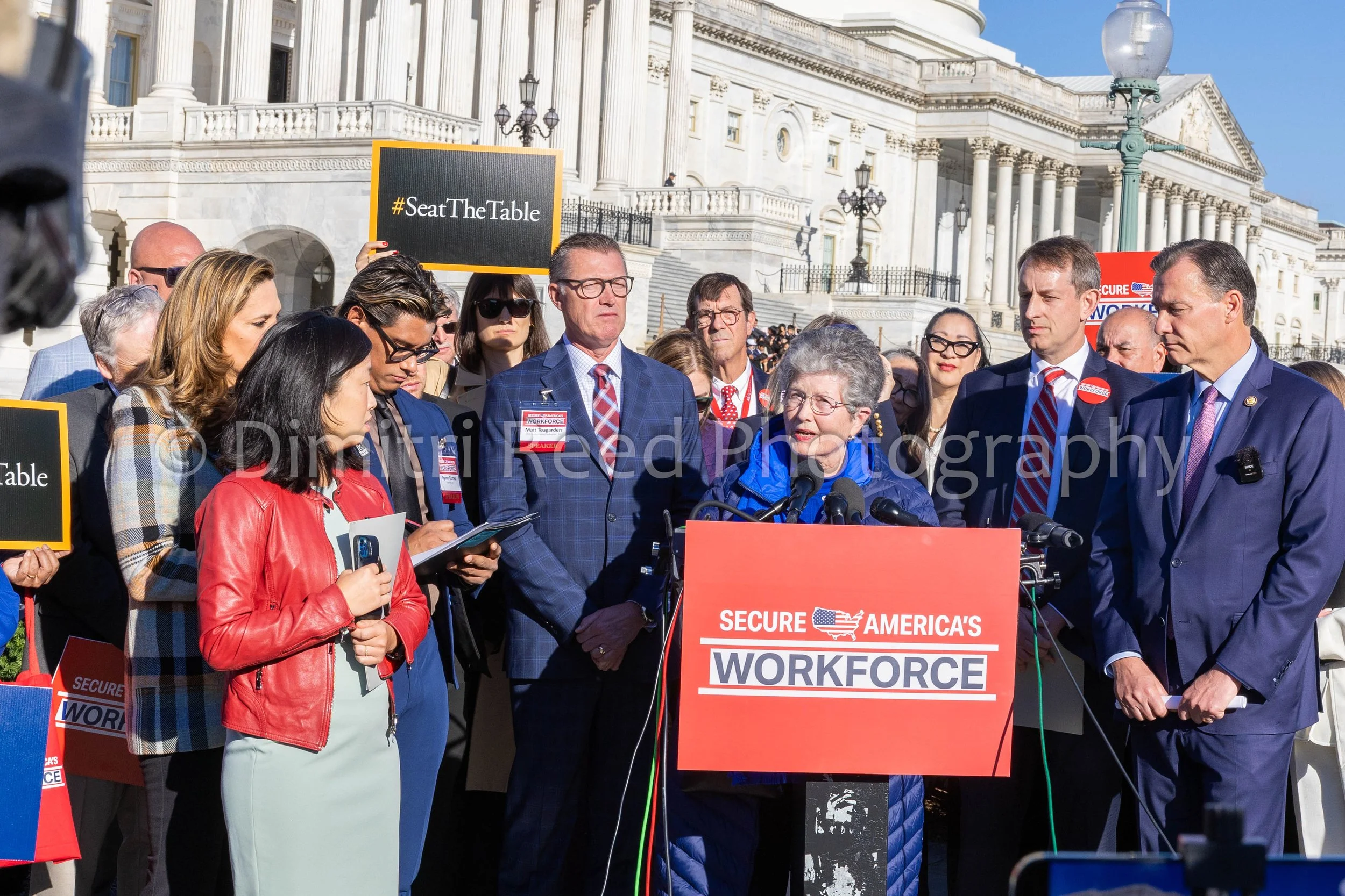 A group of people gathered outside the Capitol building during a protest or rally, holding signs and speaking at a podium that reads 'Secure America's Workforce.'