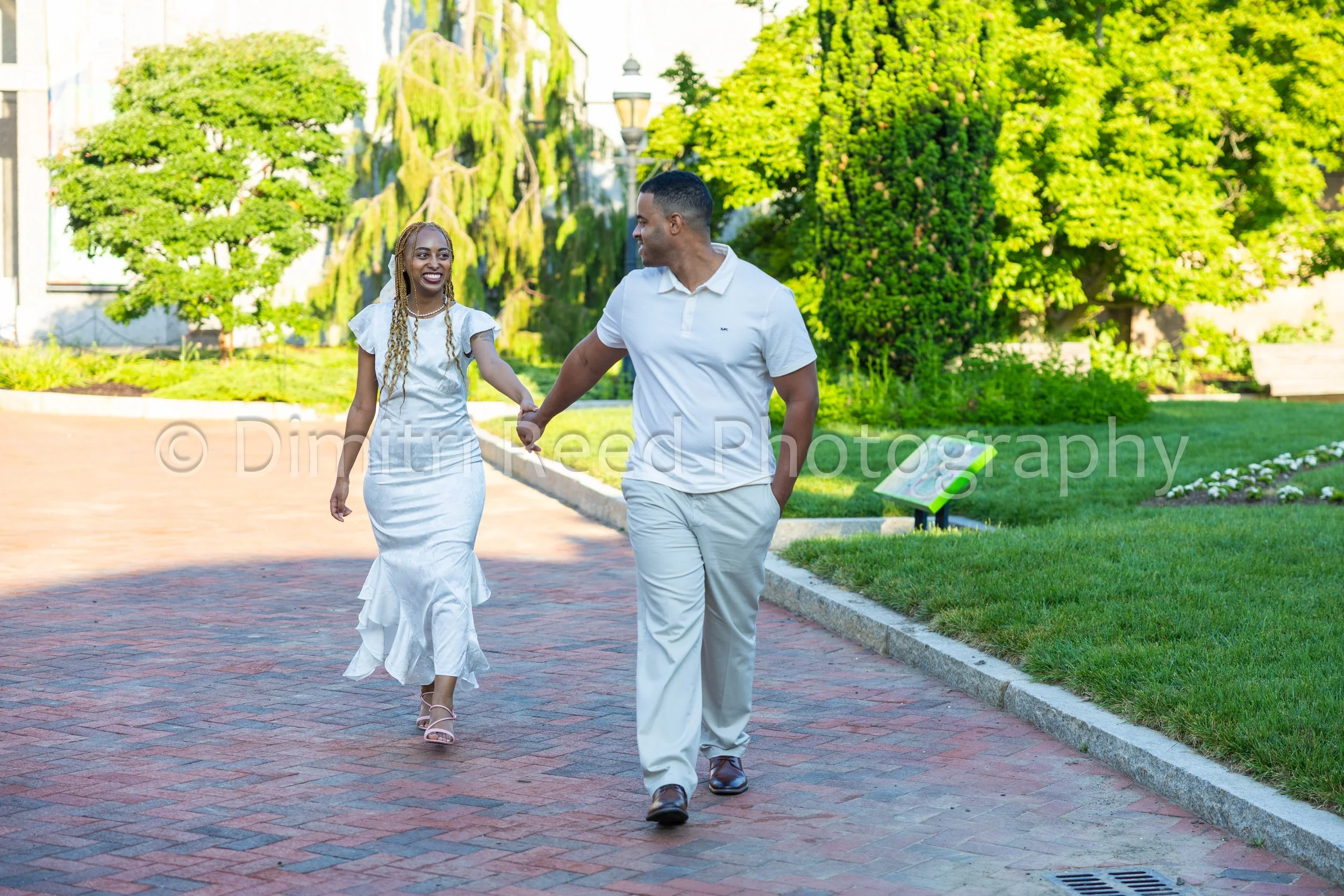 A man and woman walking hand in hand in a park, smiling at each other, with trees and greenery in the background.