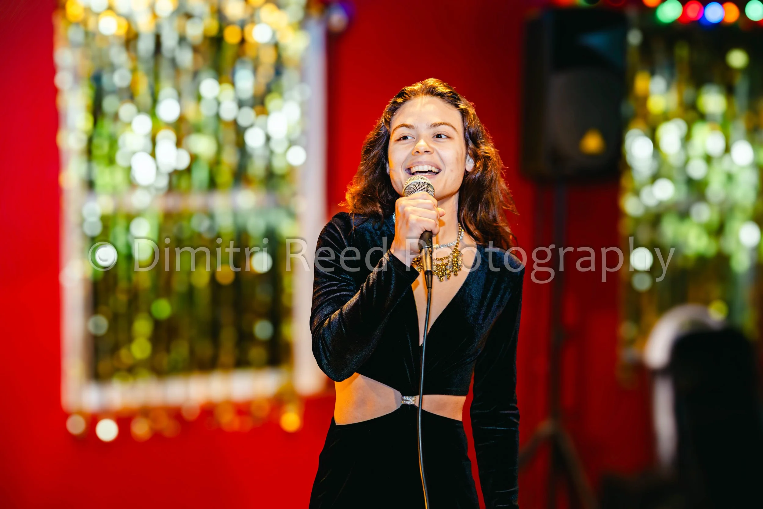 Young woman with long wavy hair holding a microphone, smiling, dressed in a black outfit, performing on stage with colorful blurred lights in the background.