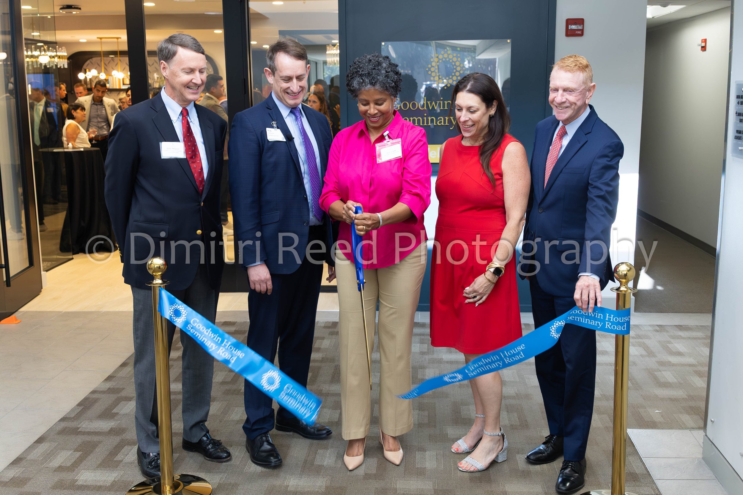 Five professionally dressed people at a ribbon-cutting ceremony in front of a sign that reads 'Goodwin House Seminar Road.' They are smiling and holding a blue ribbon, with one woman in pink holding scissors.