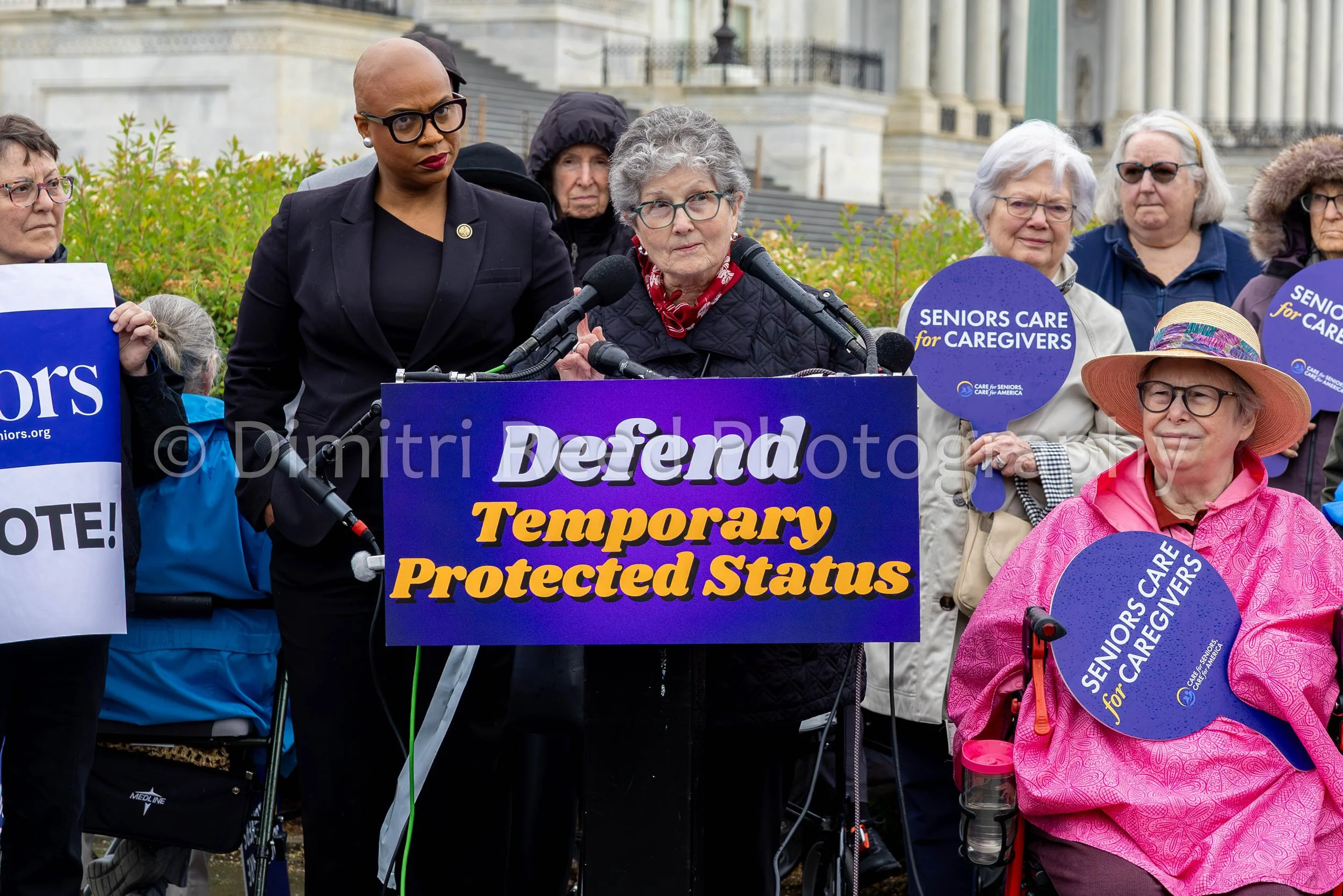 A group of elderly women and a younger woman speaking at a rally for seniors' rights, holding signs that read 'Seniors Care for Caregivers' and a purple sign that says 'Temporary Protected Status.' The women are outside a government building.
