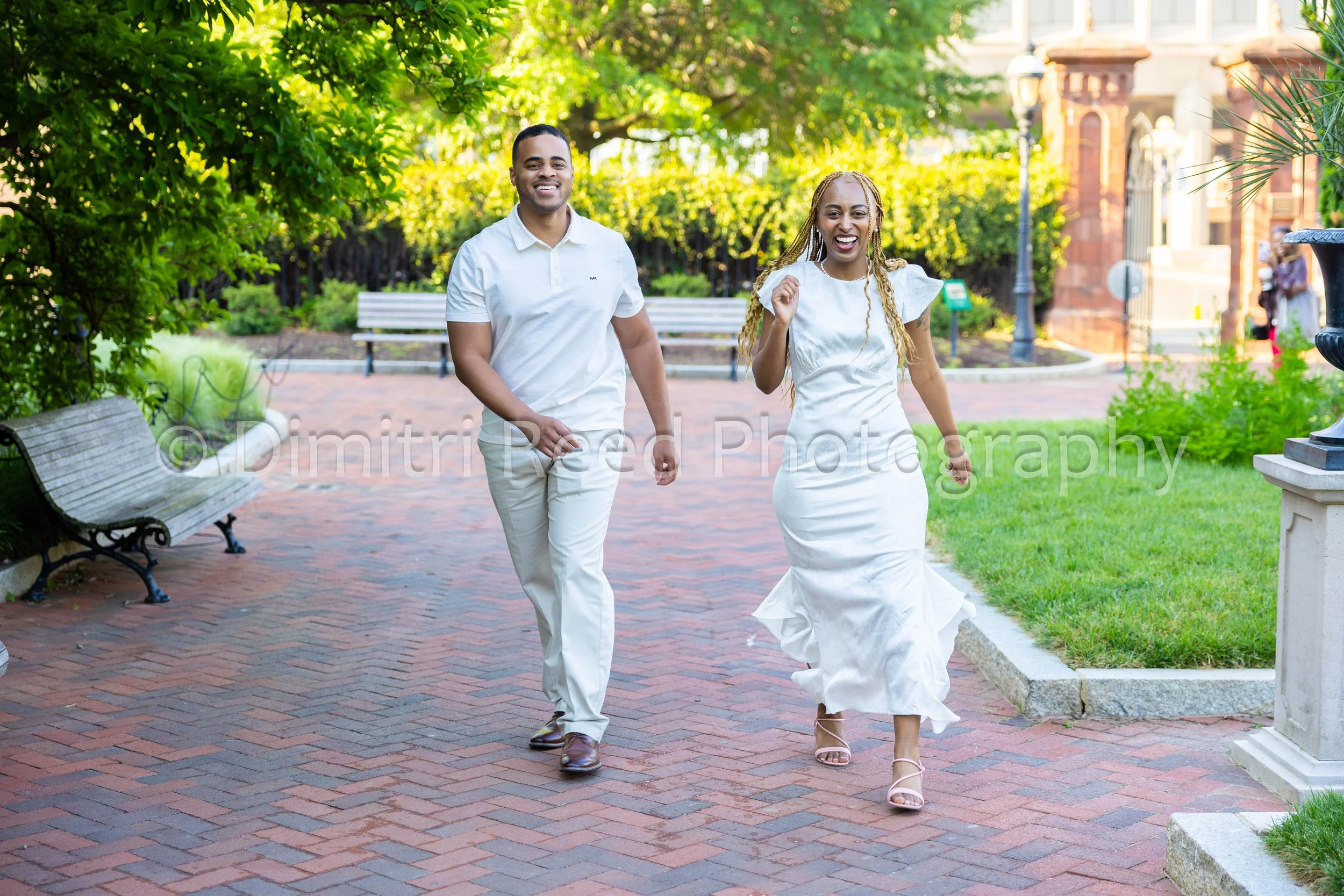A smiling man and woman walking outdoors on a sunny day, surrounded by trees and greenery. The woman is wearing a white dress and he is wearing a white shirt and light-colored pants.