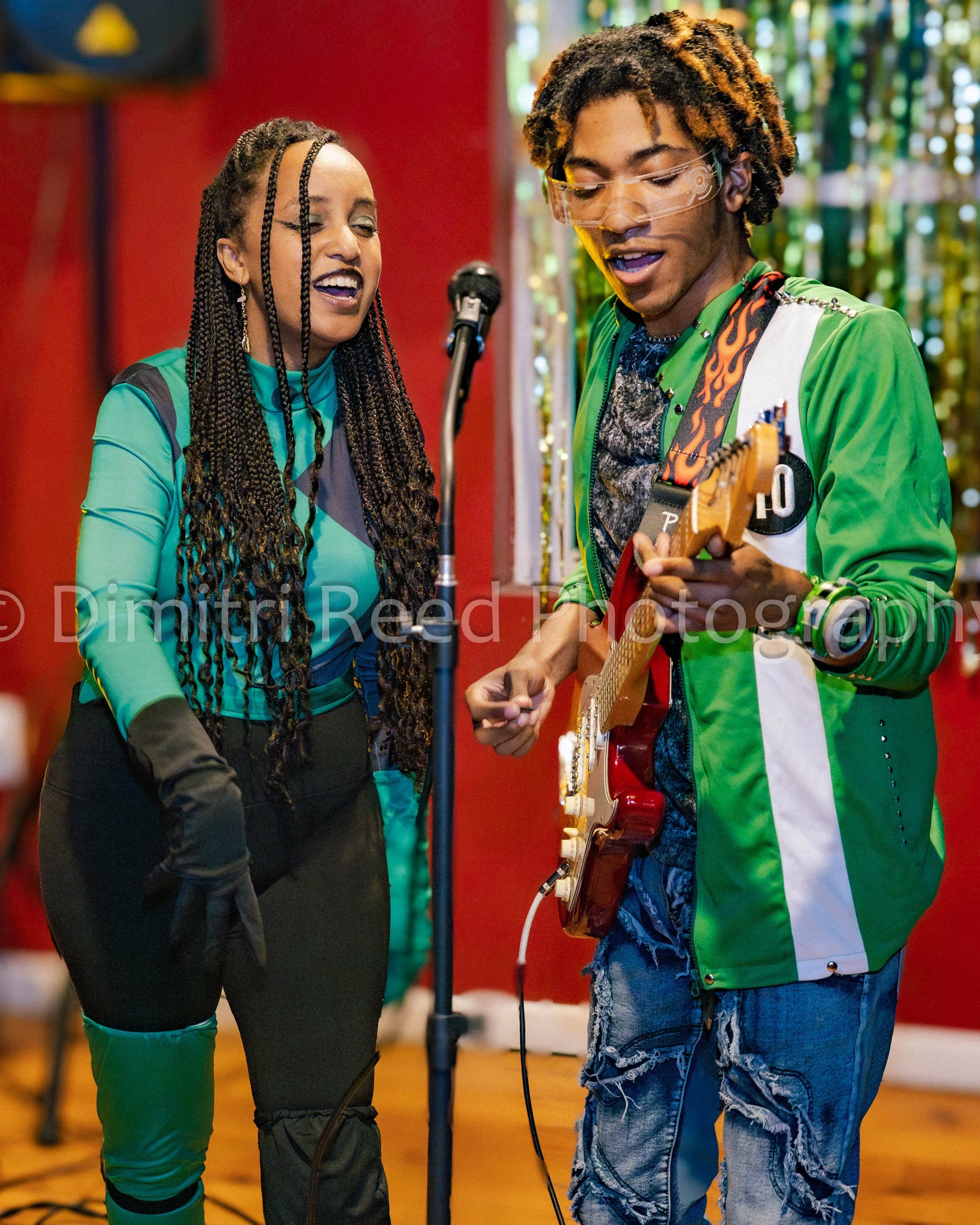 Two people, a woman and a man, singing together in a recording studio. The woman has long braided hair and is wearing teal and black clothing. The man has dreadlocks, is wearing glasses, a green and white jacket, and playing an electric guitar. They 
