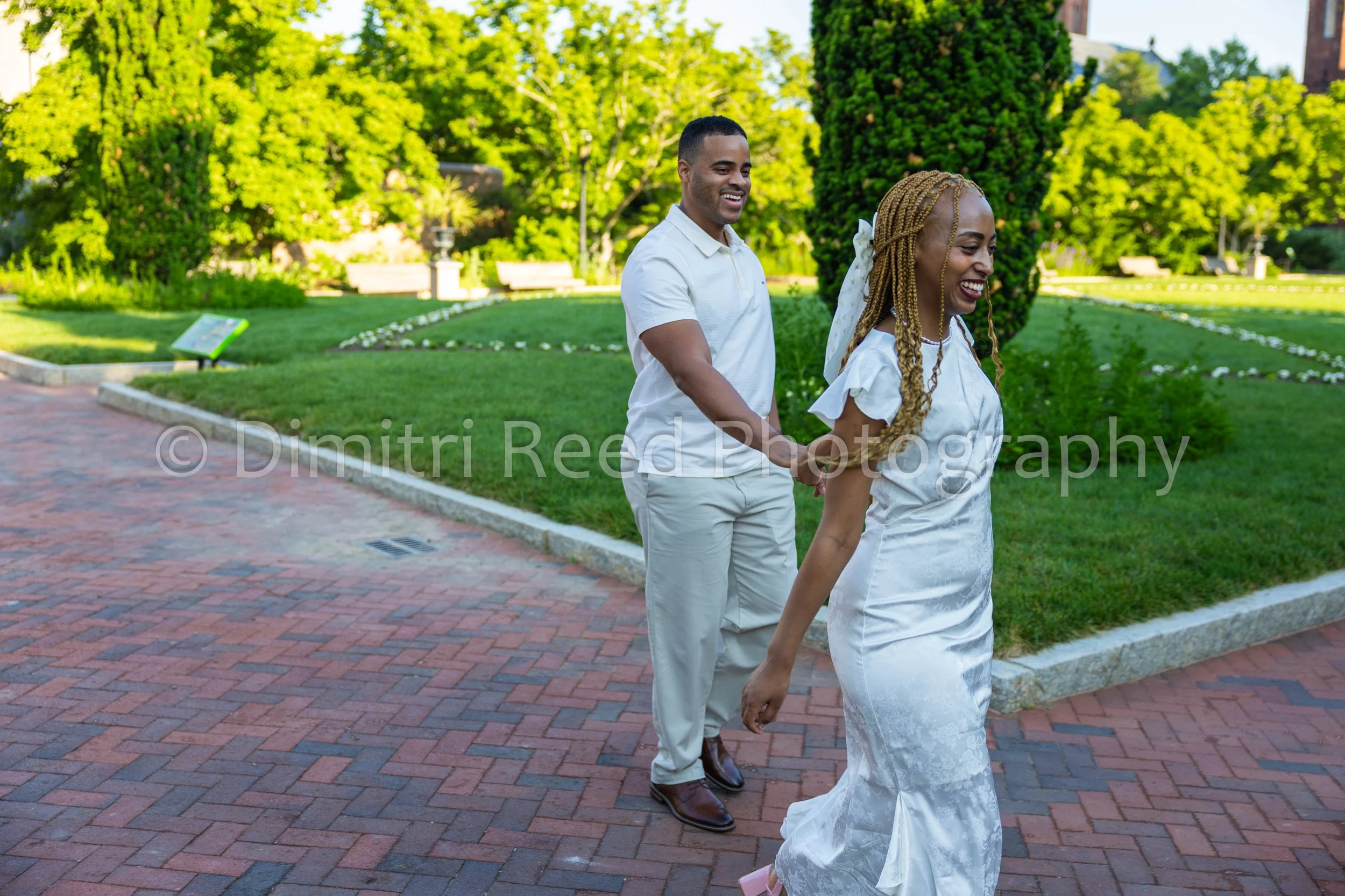 A happy couple dressed in white holding hands and walking together in a park on a sunny day, with trees and benches in the background.