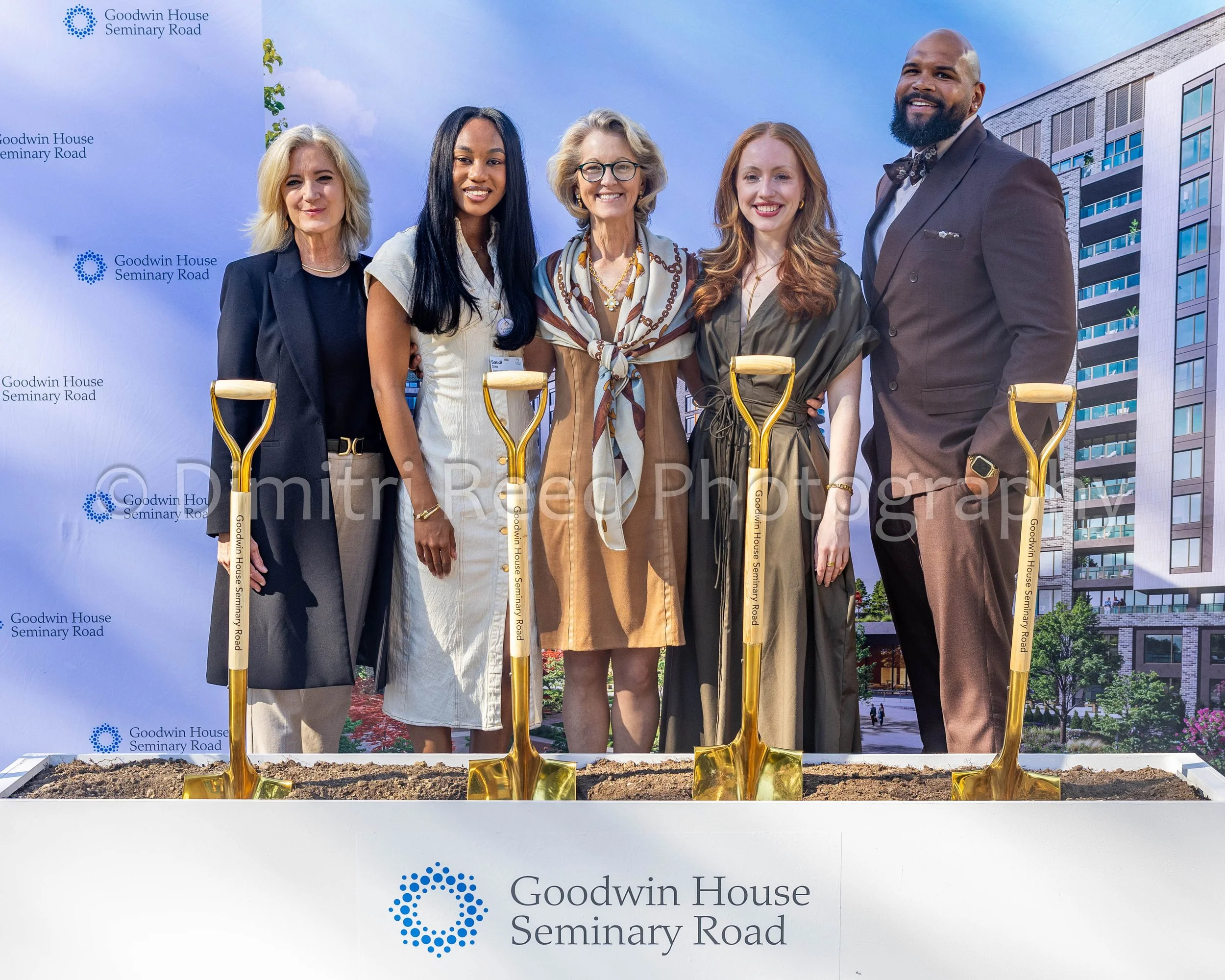 A group of six diverse people standing together outdoors on a bright day, smiling, behind a white backdrop with the logo and name of Goodwin House Seminary Road. The group appears to be at a groundbreaking event, with shovels in front of them and a modern building visible in the background.
