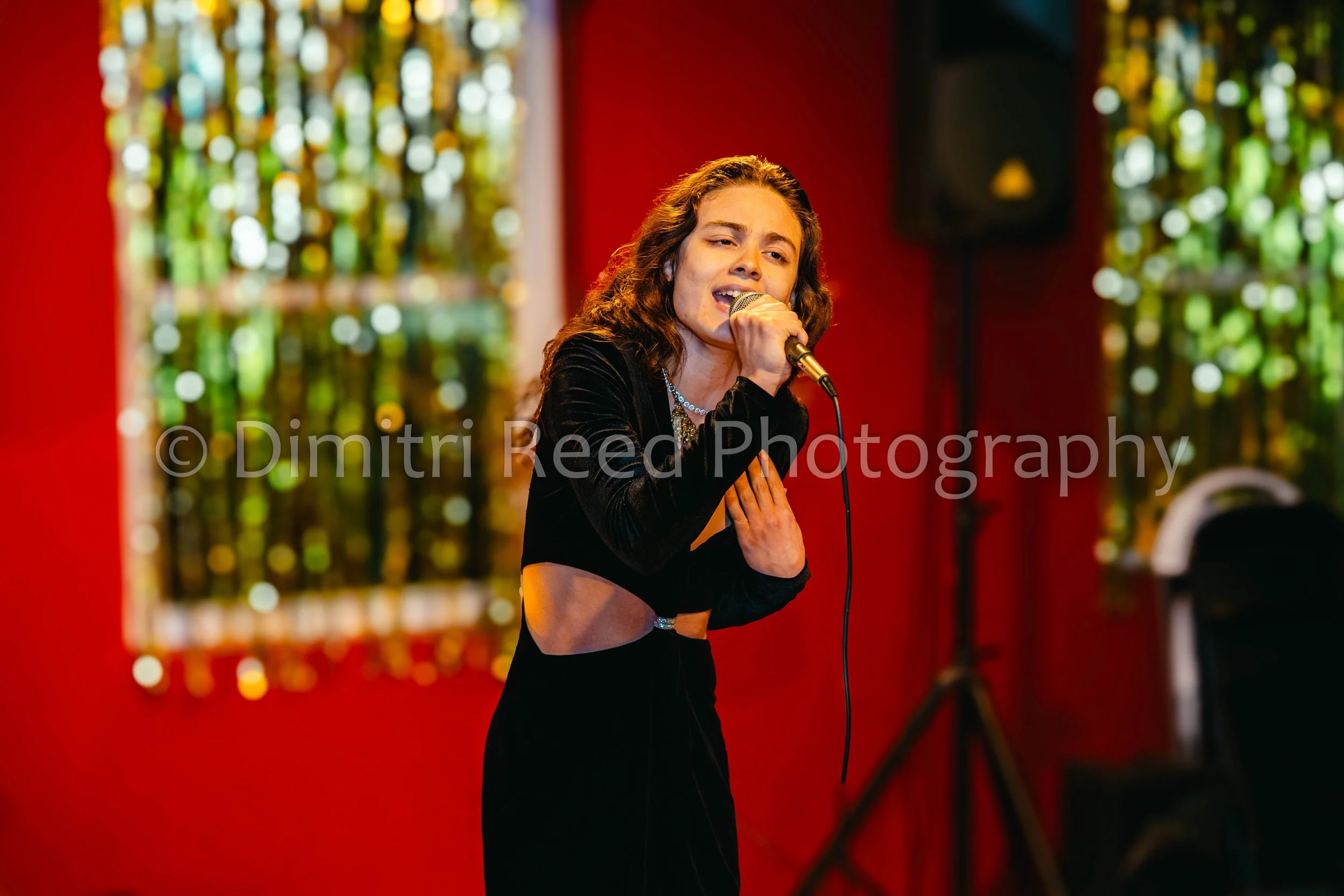 A young woman with curly hair singing into a microphone on stage, wearing a black top with cutouts, against a blurred background of colorful lights and red wall.
