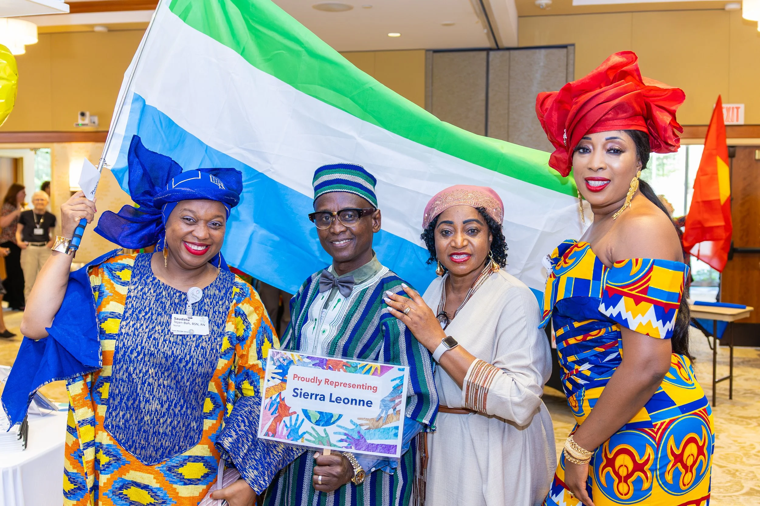 Group of four people dressed in colorful traditional African attire posing at an indoor event, holding a Sierra Leone flag and a sign that reads 'Proudly representing Sierra Leone.' There are other attendees in the background.