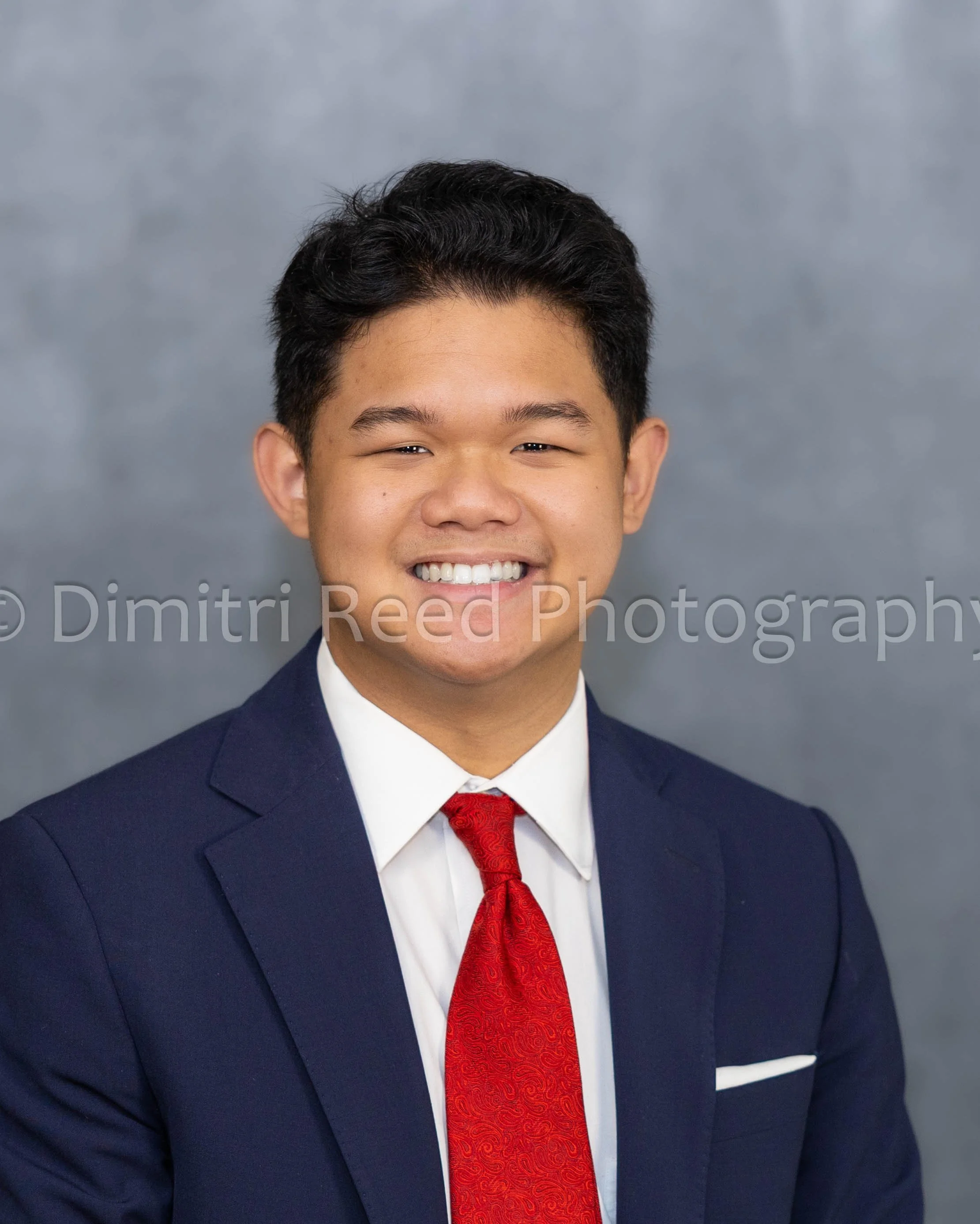 A young man in a navy suit, white shirt, and red tie smiling in front of a gray background.
