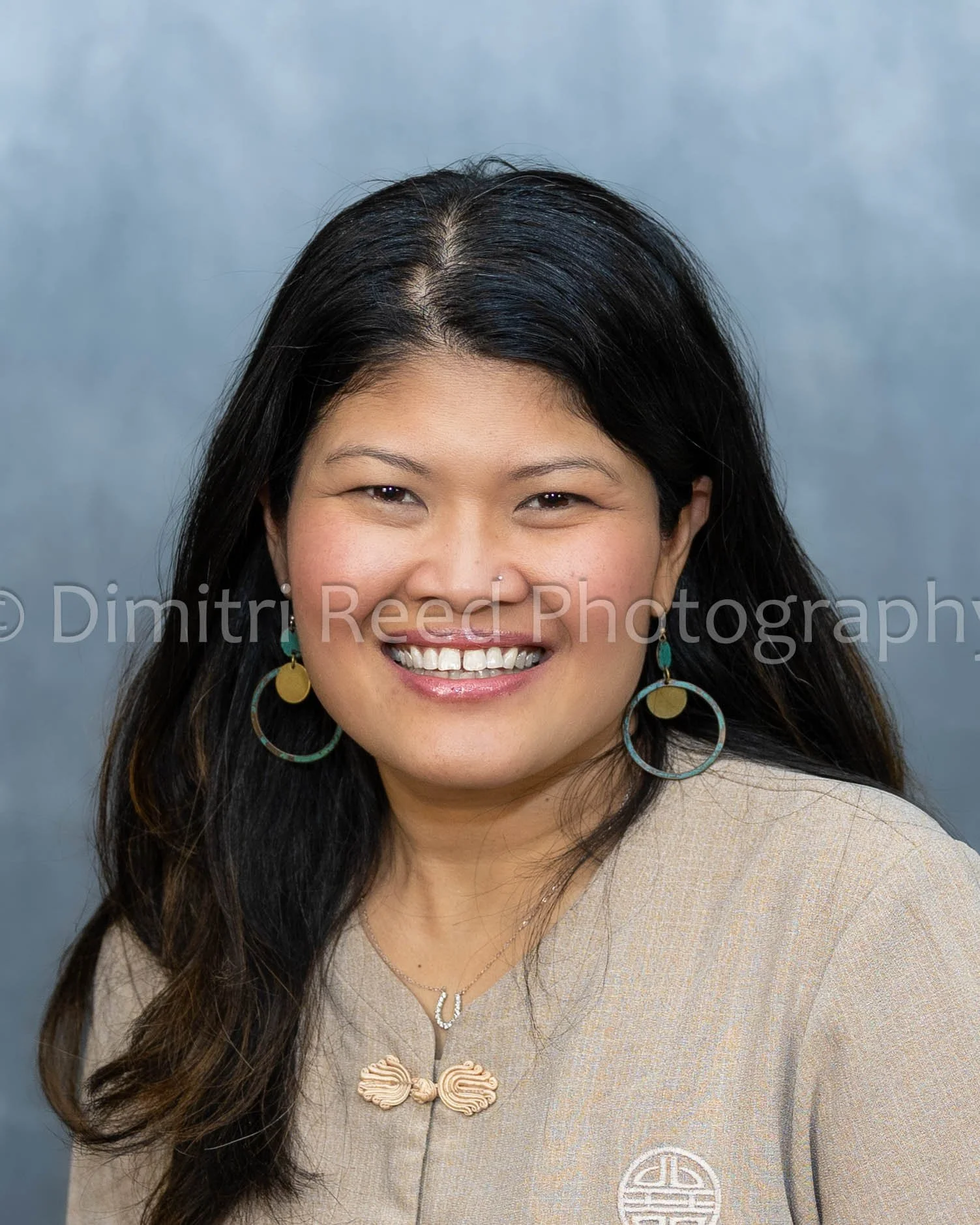 Portrait of an Asian woman with long dark hair, smiling, wearing colorful earrings, a beige traditional outfit, and a necklace, against a gray background.