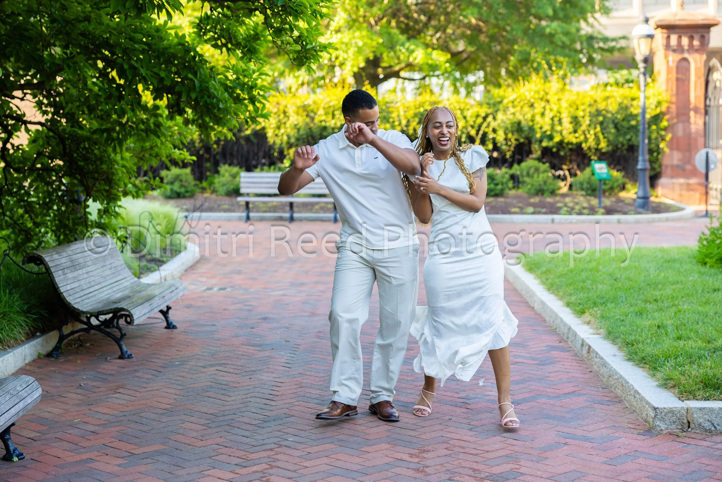 A smiling couple dancing and enjoying themselves outdoors on a brick path in a park with lush green trees and benches.