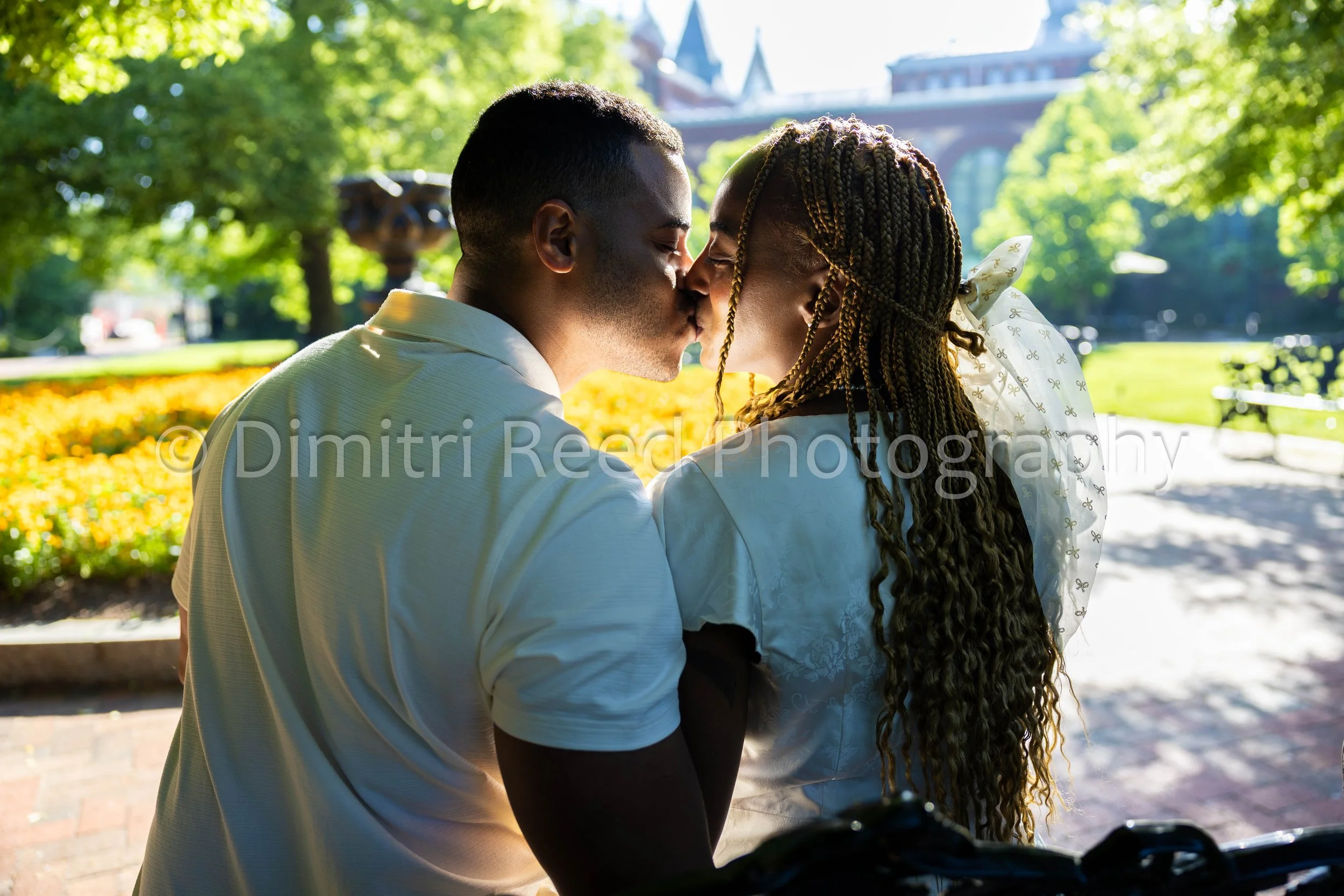 A couple kissing outdoors in a park with green trees and a historic building in the background on a sunny day.