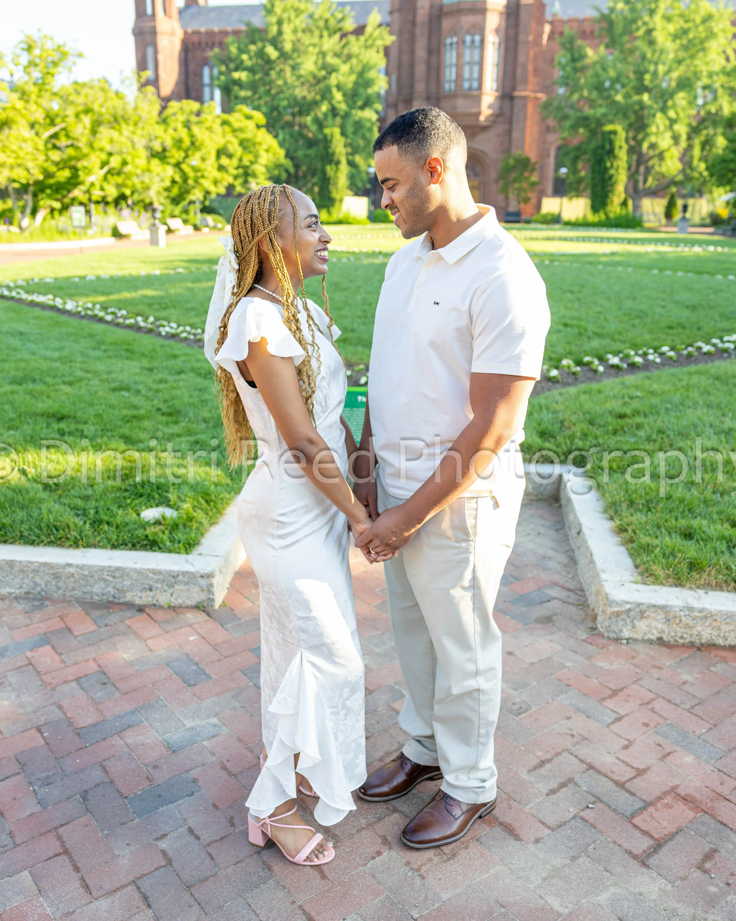 A couple dressed in white, holding hands and smiling at each other, standing on a brick path in a park with greenery and a red brick building in the background.