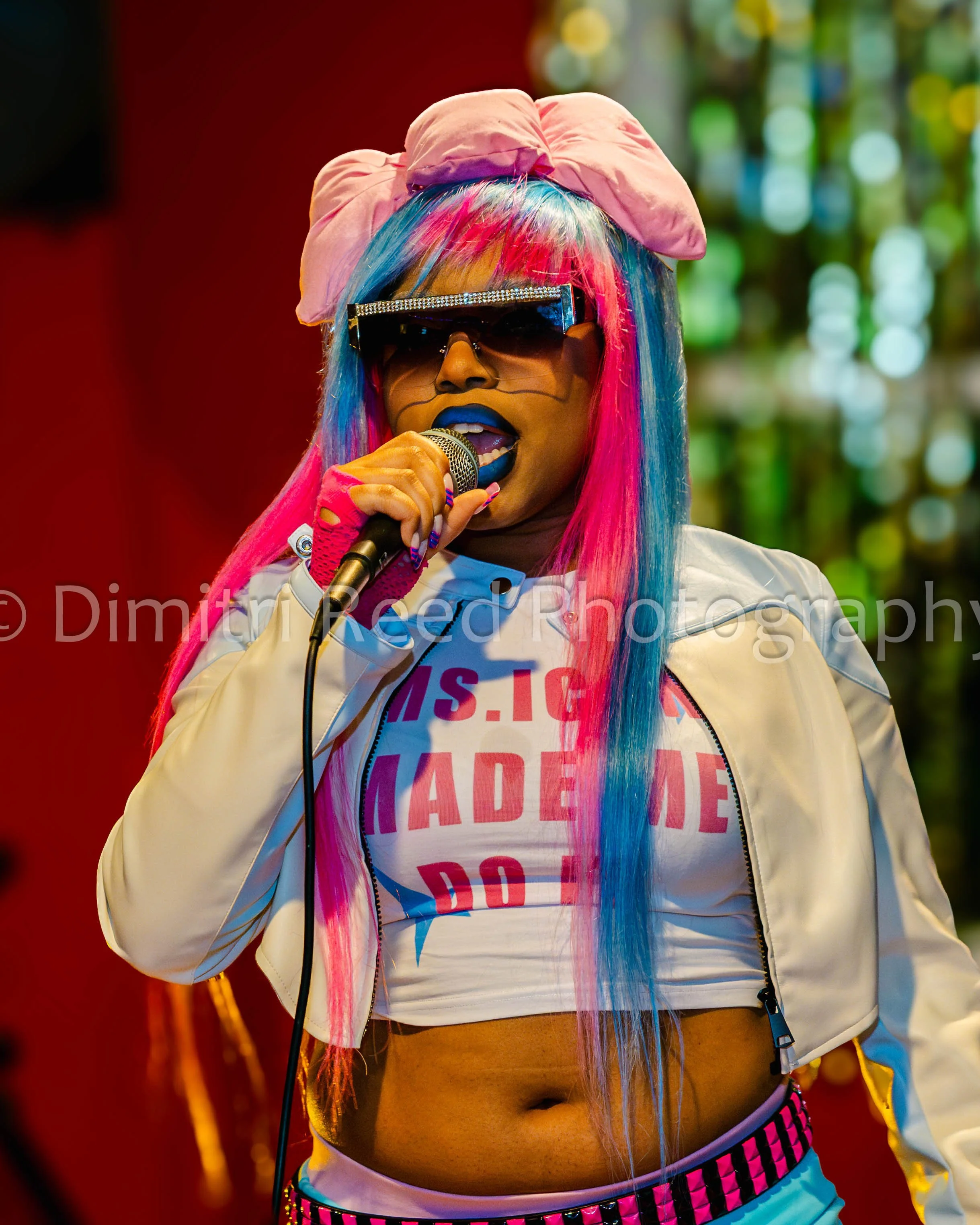 A person with colorful hair wearing large sunglasses, a pink headscarf, a white jacket, a graphic T-shirt, and shorts. They are holding a microphone and appear to be performing on stage.