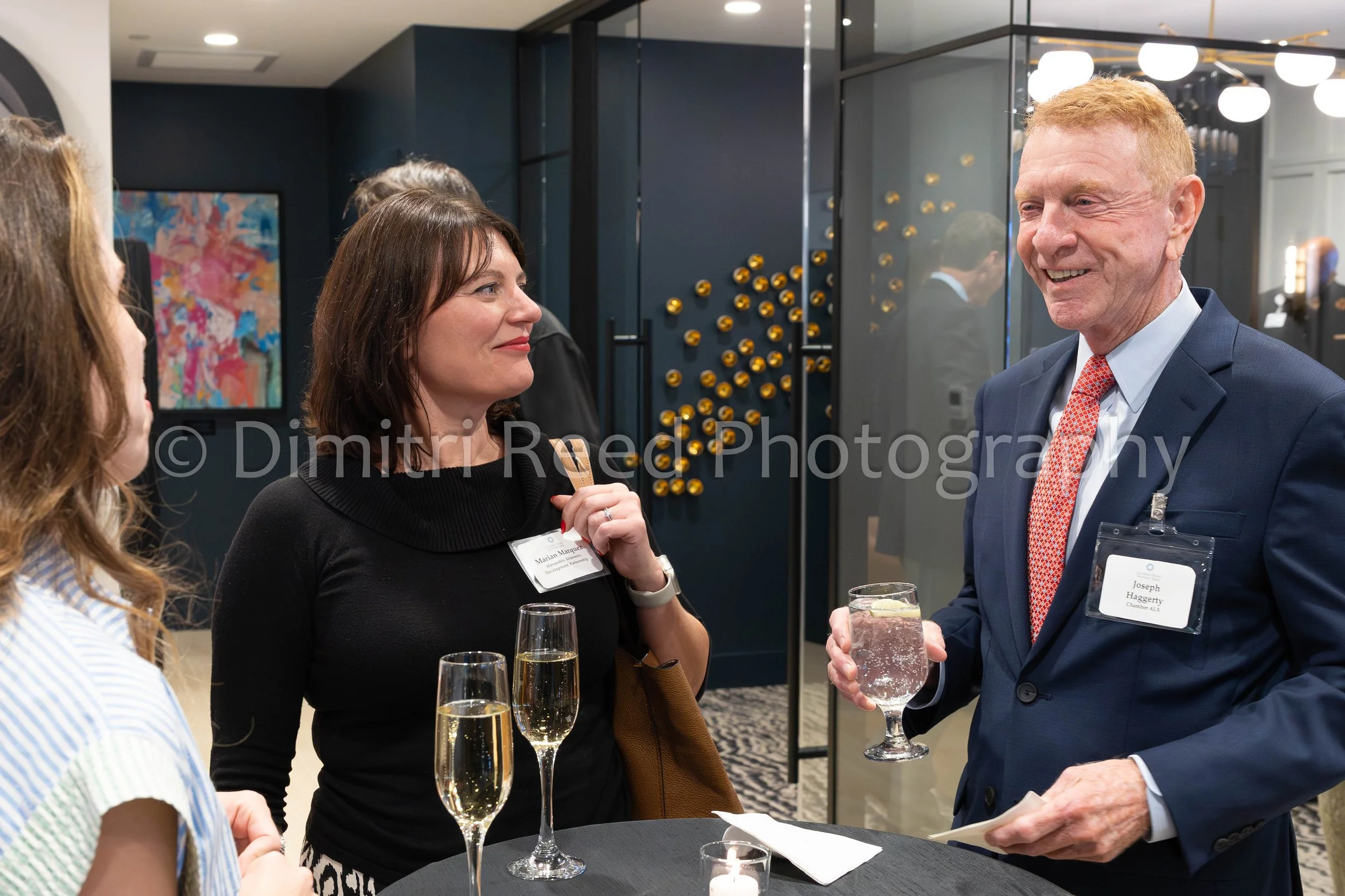 A group of people at a networking event, including a woman named Marian Marquez and a man named Joseph Haggerty, engaged in conversation, holding drinks in a modern, upscale indoor setting with dim lighting and decorative wall accents.