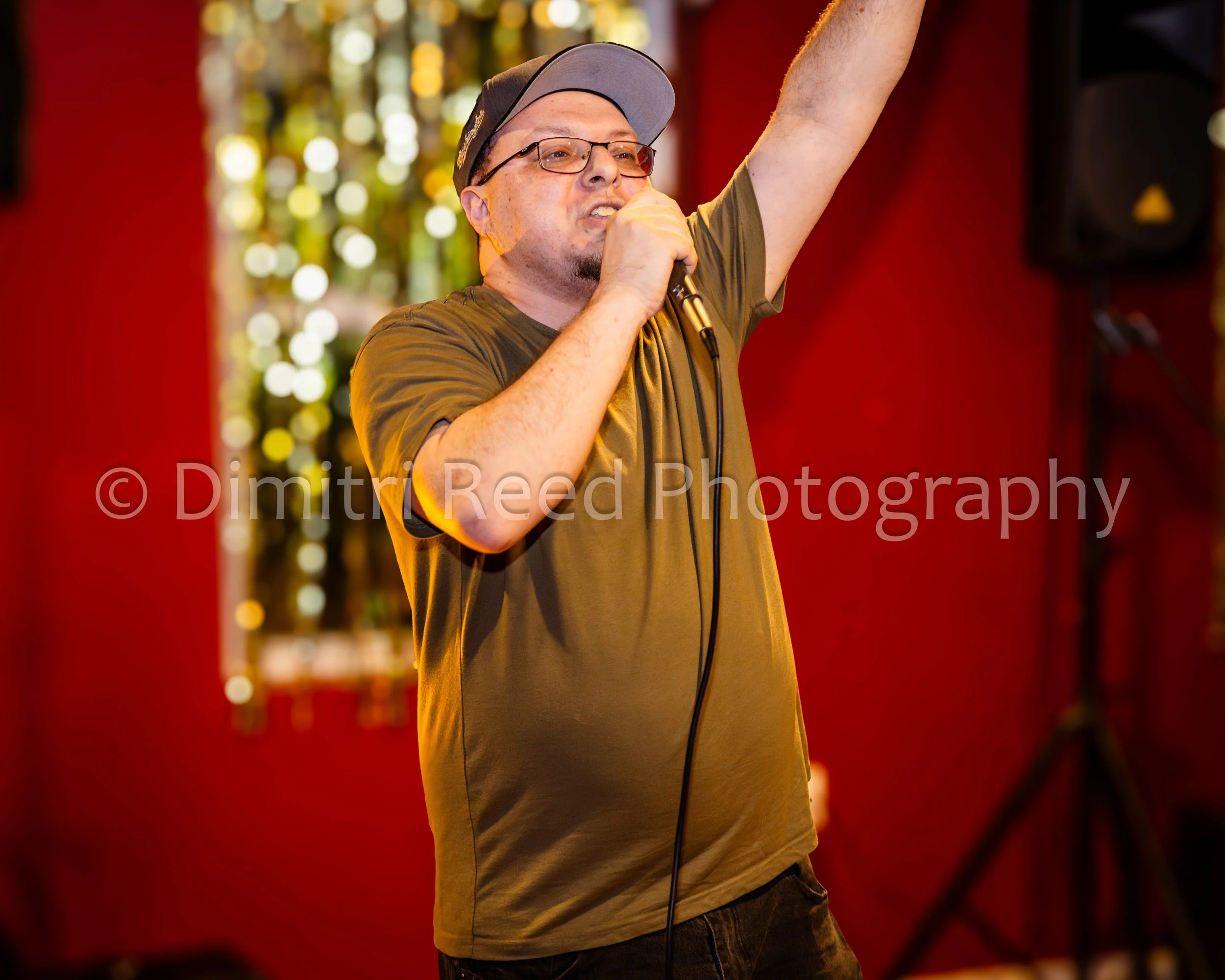 A man wearing a gray cap and glasses singing or speaking into a microphone with his right arm raised, against a red and yellow blurred background.