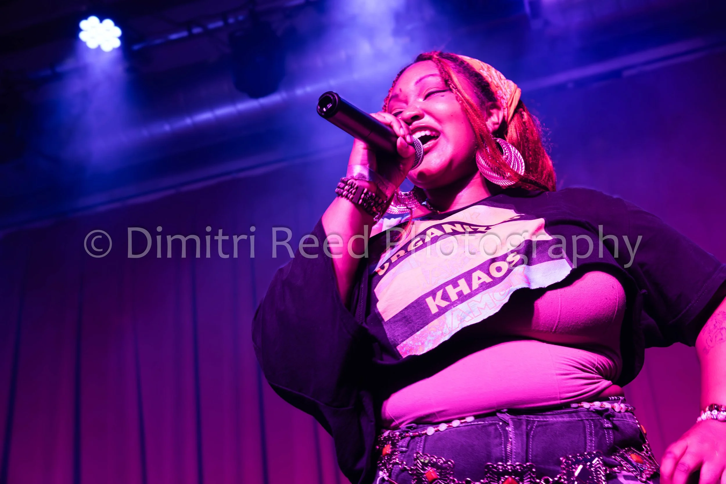A woman with red braided hair singing into a microphone on stage with purple lighting, wearing large earrings, a graphic t-shirt, and a black jacket.