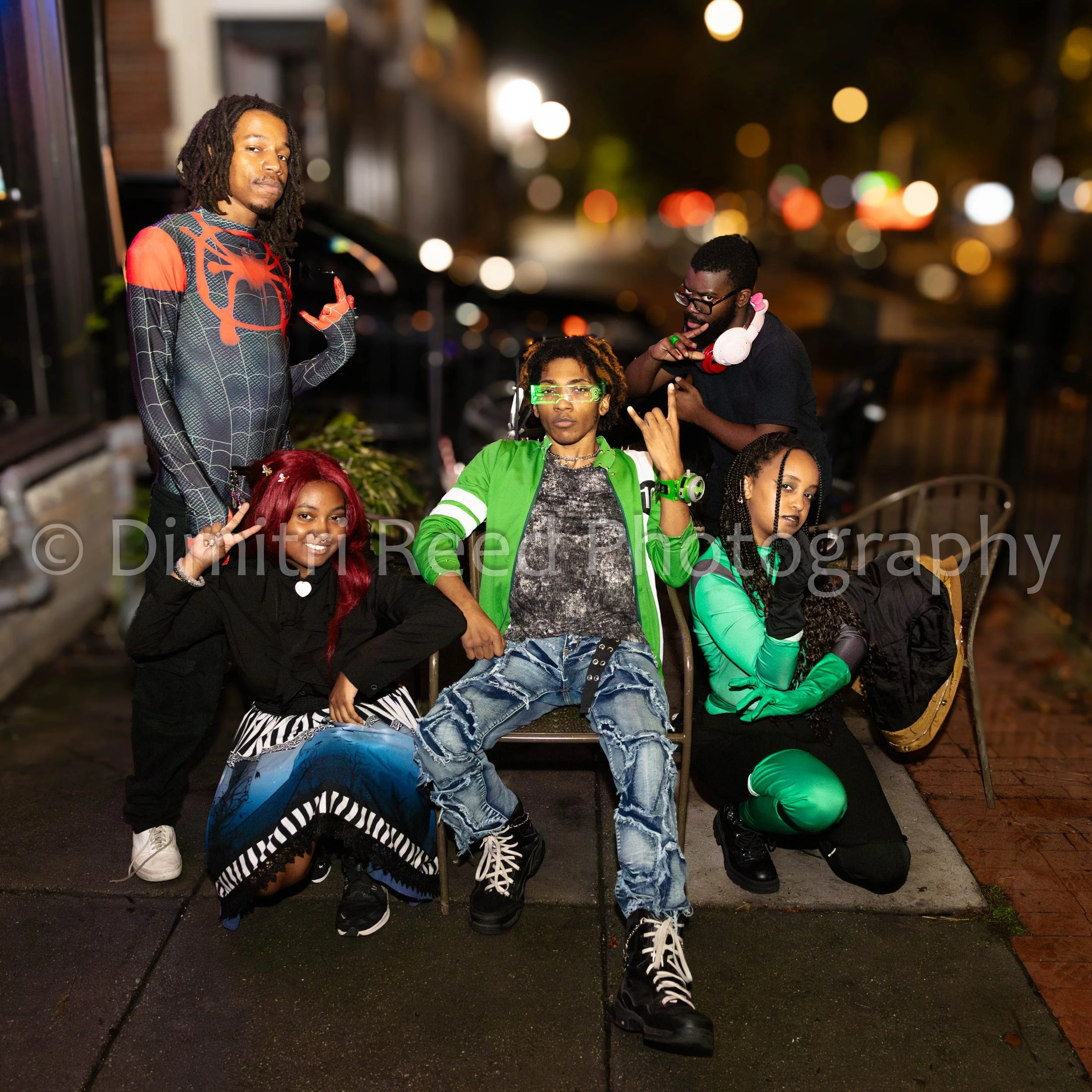 A group of five young adults posing on a city street at night. They are dressed in colorful and trendy clothing, with some wearing glasses and headphones. The background features blurred city lights and a sidewalk, creating a lively urban atmosphere.