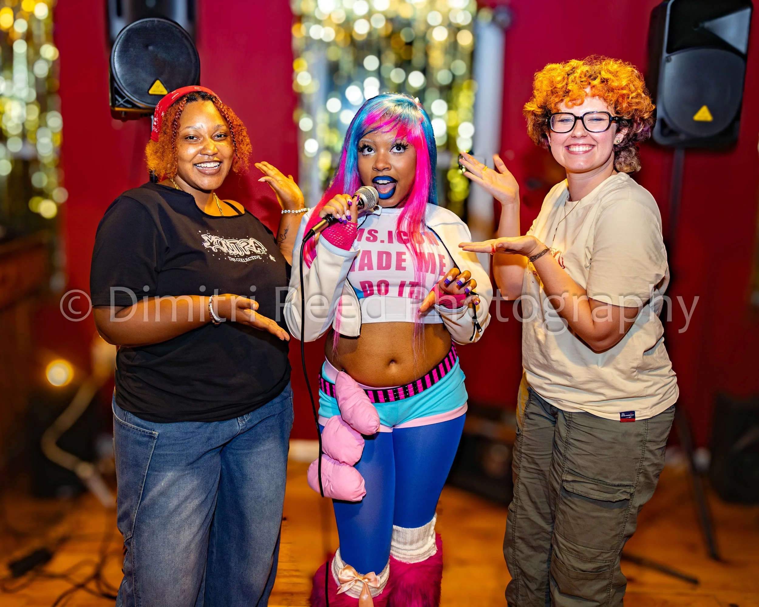 Three women and a girl posing together at a holiday event. The girl is dressed in a colorful costume with a pink and blue wig and pink gloves, holding a microphone. One woman is wearing glasses and a beige shirt, and another woman is wearing a black 