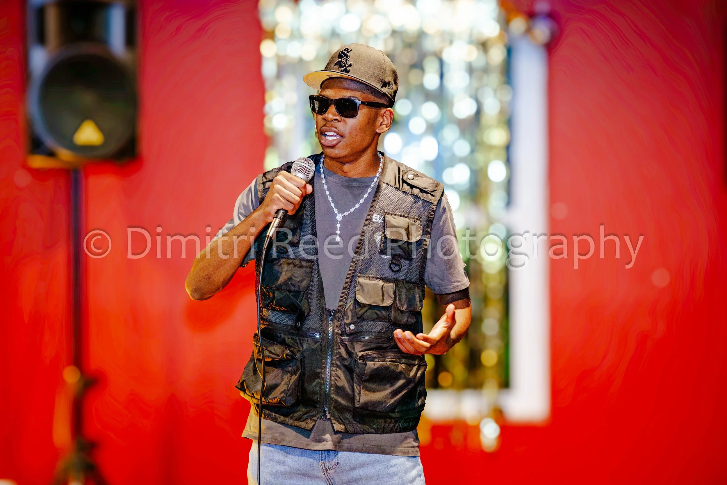 A young man wearing sunglasses and a cap, holding a microphone, stands in front of a red backdrop with blurred bokeh lights.