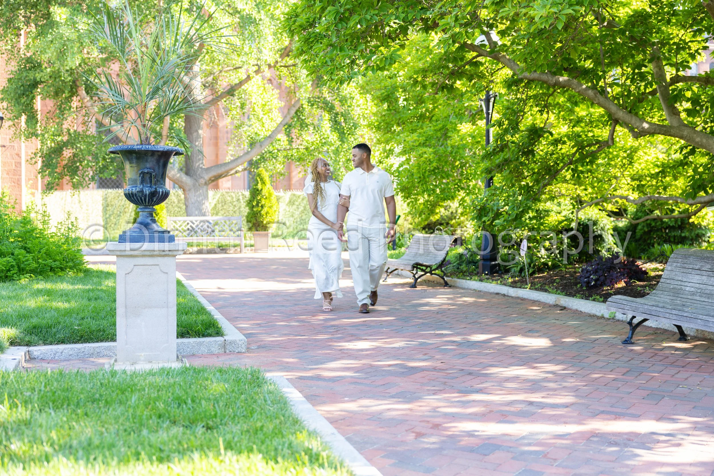 A couple walks hand in hand through a lush green park with large trees, benches, and a decorative blue urn with tall grass.