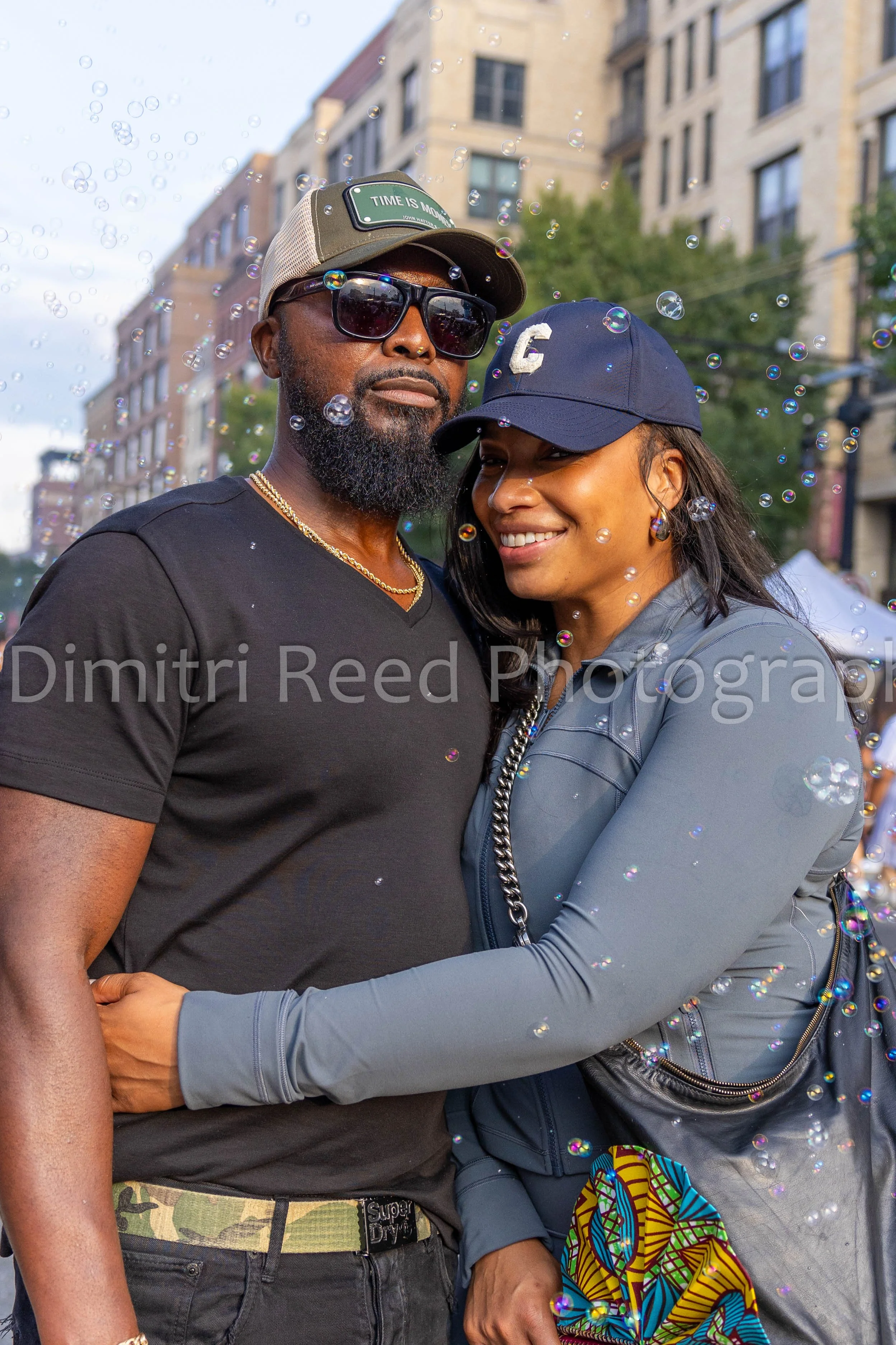 A man and woman are standing close together outdoors, smiling and embracing. They are surrounded by floating bubbles with an urban background of apartment buildings and trees.