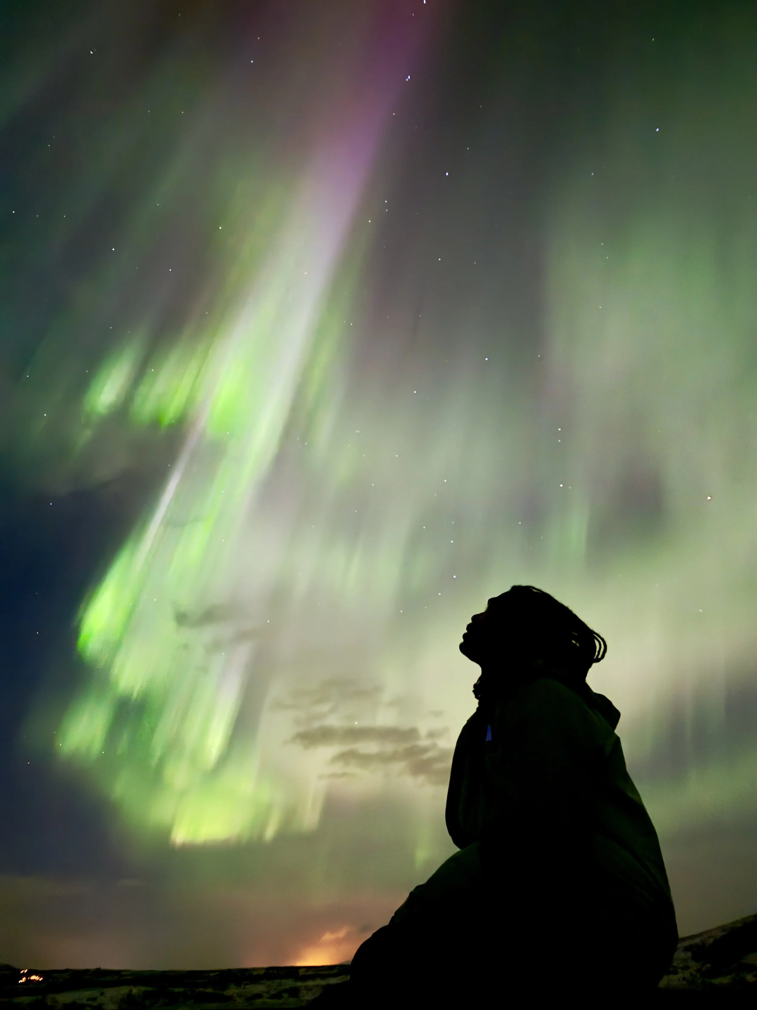 Silhouette of a person standing outside at night, watching the Northern Lights (aurora borealis) in the sky, with stars visible in the background.