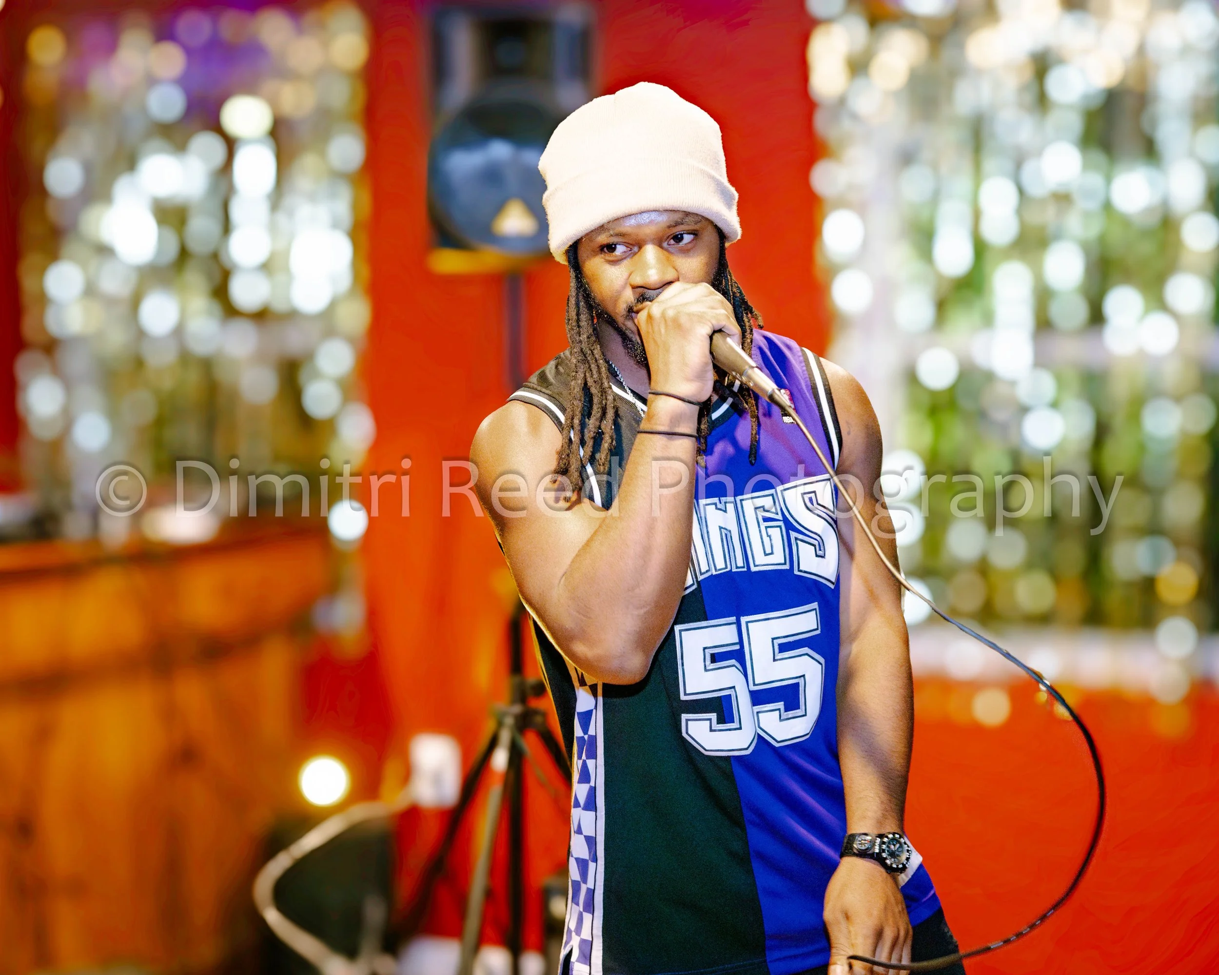 A man in a basketball jersey and a white beanie holding a microphone in an indoor setting with colorful bokeh lights in the background.
