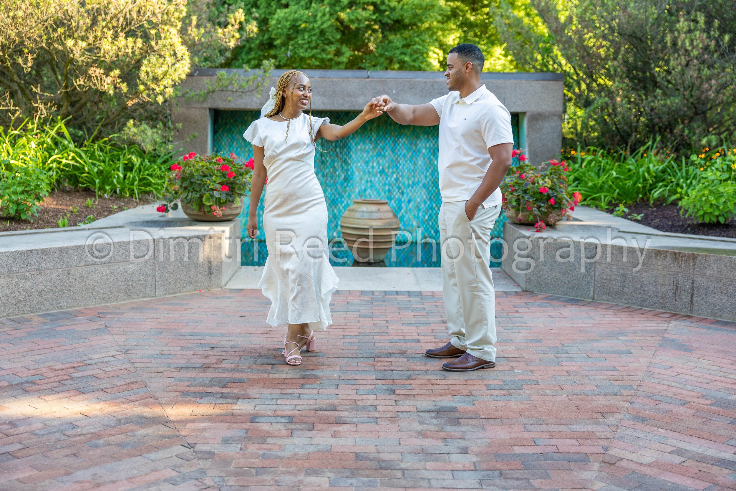 A couple dance hand in hand outdoors on a brick paved area surrounded by greenery, with the woman wearing a white dress and heels and the man dressed in a white polo and beige pants.