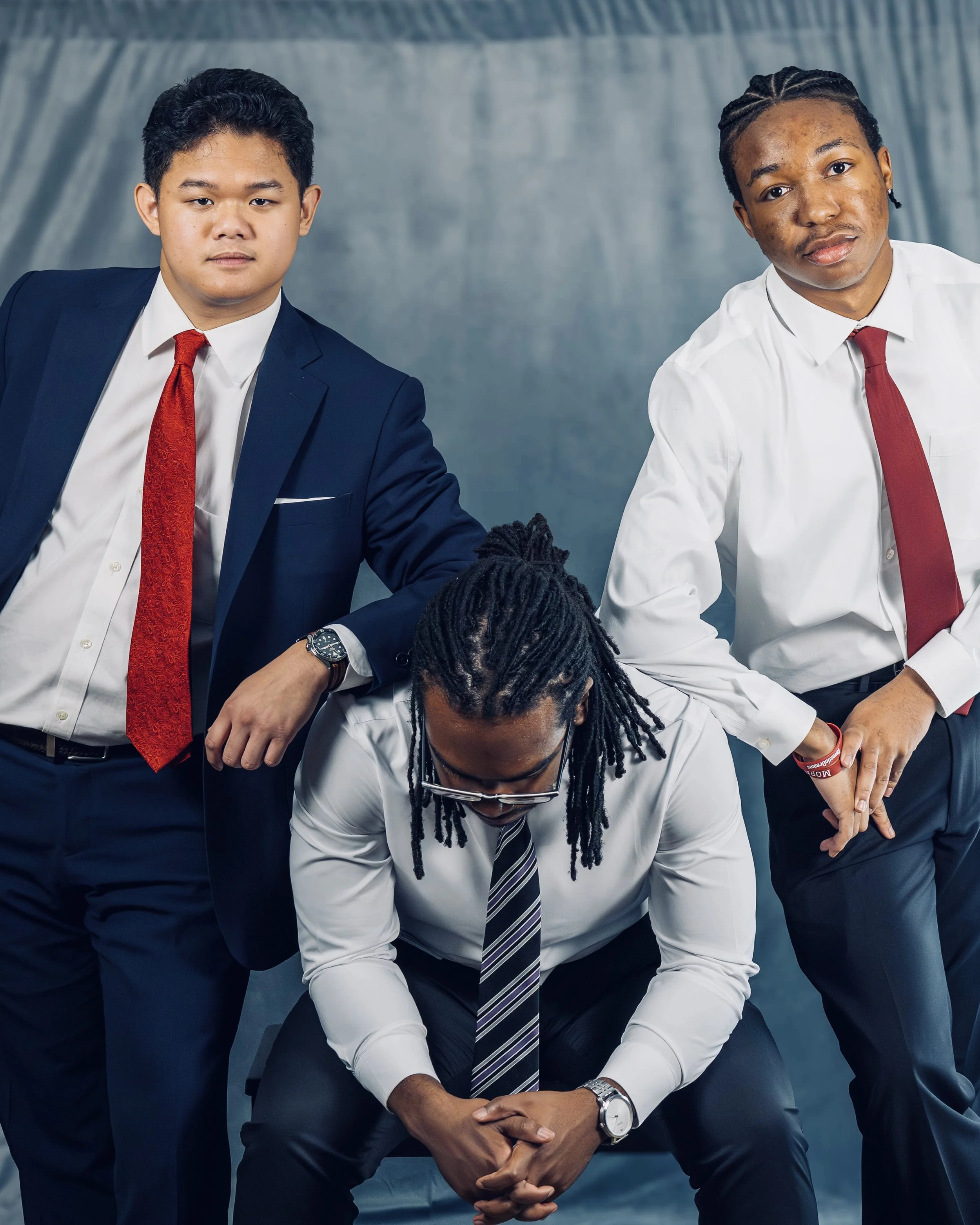 Two businessmen in formal suits with red ties pose confidently, holding hands over a person's head, in front of a plain gray backdrop.